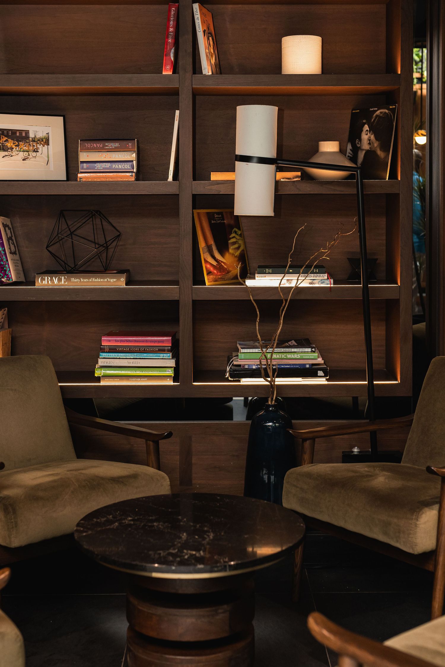 Cozy reading nook with velvet armchairs, marble side table, floor lamp, and dark wood bookshelf filled with books.
