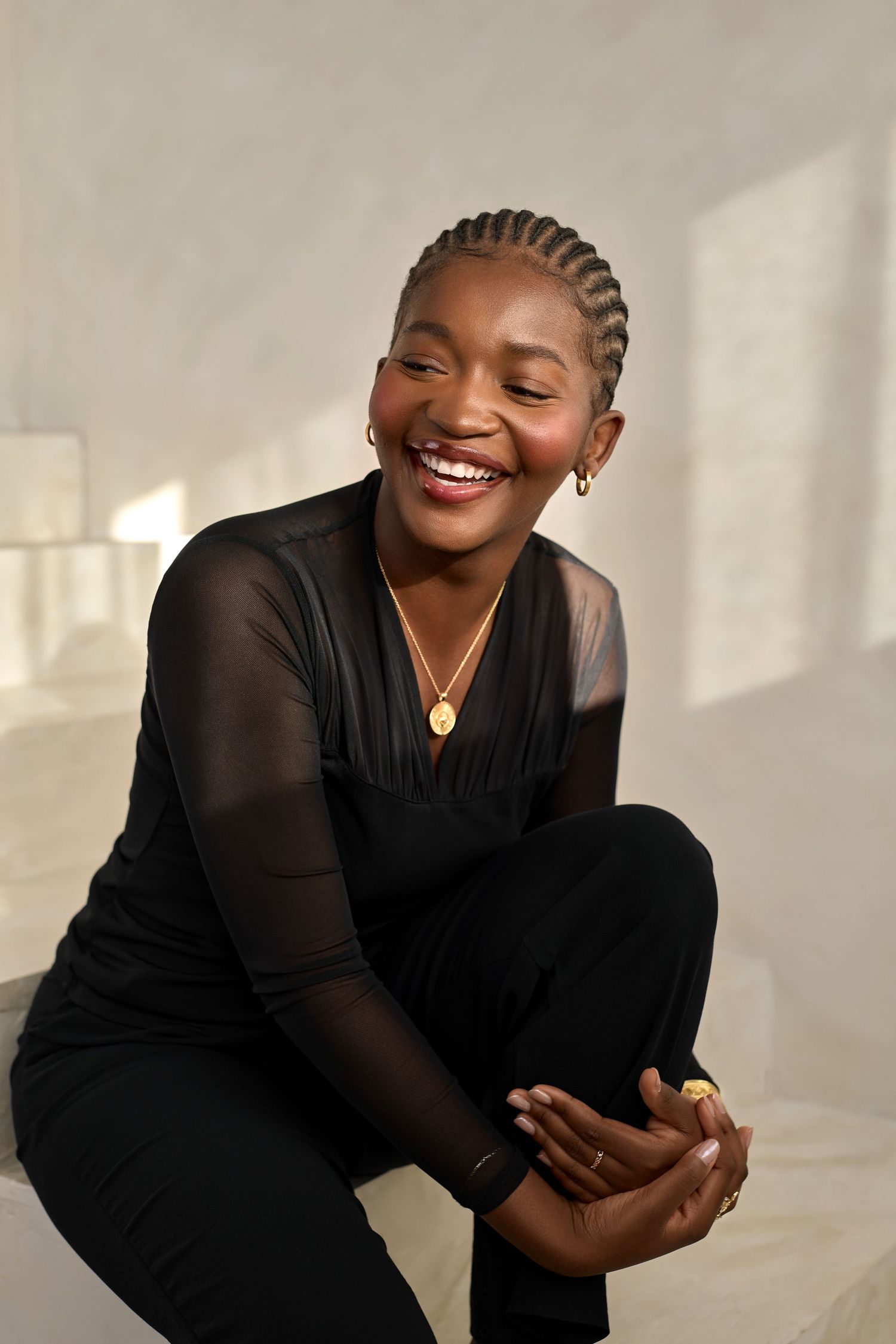 A person wearing a black sheer-sleeved dress smiles joyfully while posing in a bright studio setting.