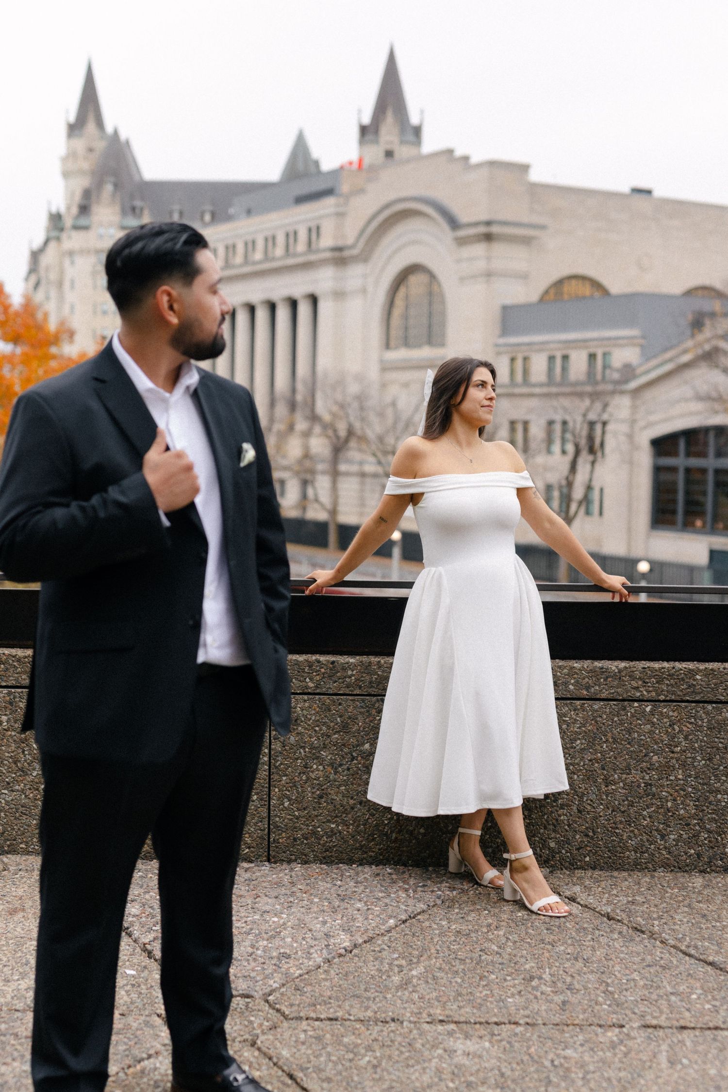A couple in formal wedding attire poses on a stone balcony with architectural buildings visible in the background.