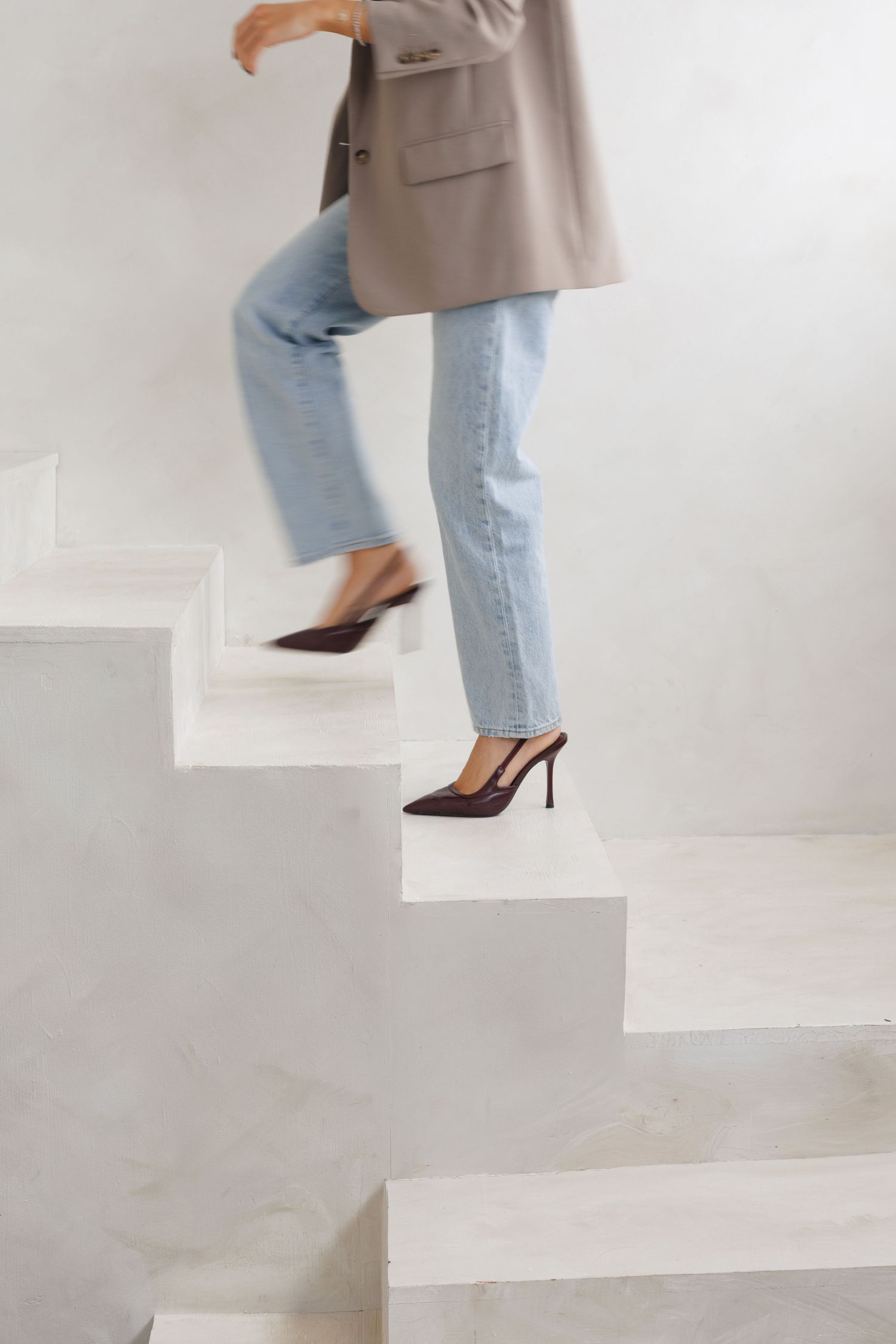 Woman in light blue jeans and dark heels ascending white stairs wearing a beige blazer.