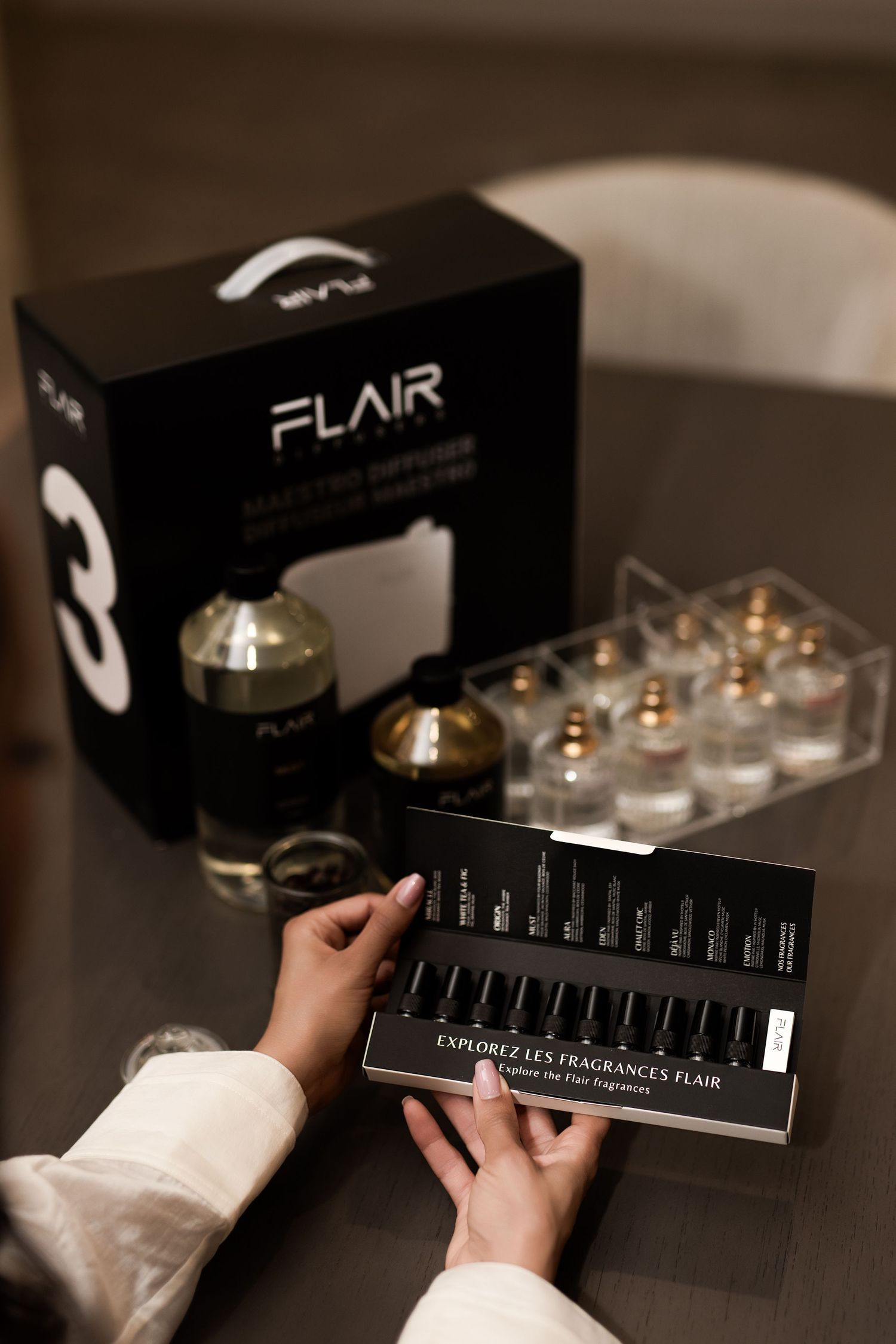 Hands holding a Flair fragrance sample card with perfume bottles and branded box on a wooden table.