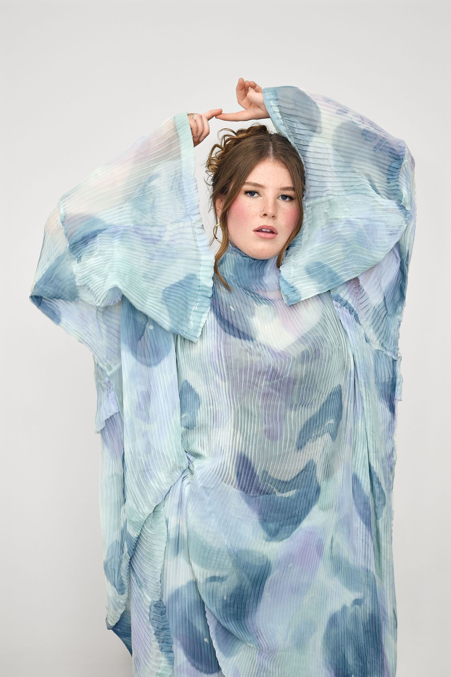Woman posing in flowing blue watercolor-print pleated sheer dress with dramatic wide sleeves against white background.