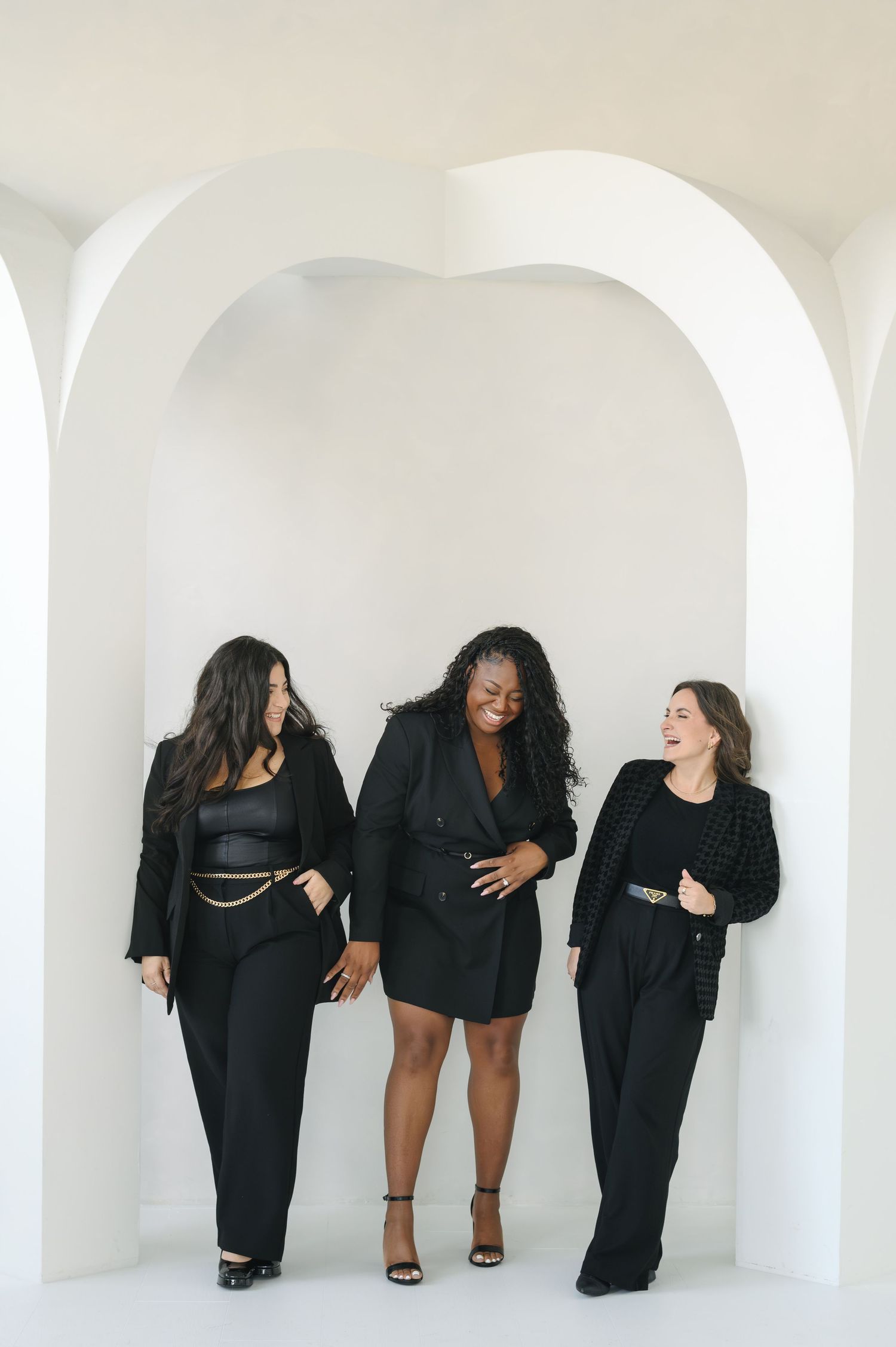 Three women in black outfits laughing together while posing in a white arched space.