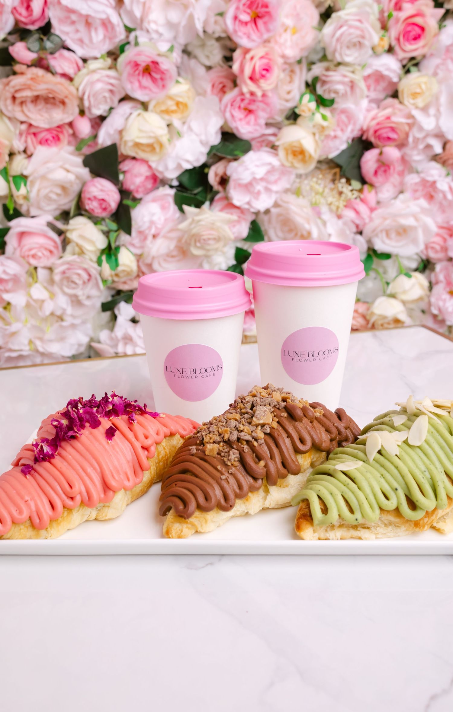 Colorful eclairs with pink, chocolate and matcha toppings displayed against a floral rose backdrop with pink coffee cups.