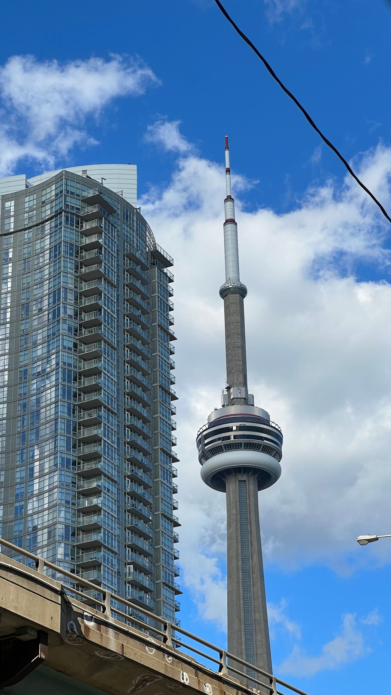The CN Tower in Toronto rises against a blue sky with white clouds, with modern glass condominiums in the foreground.