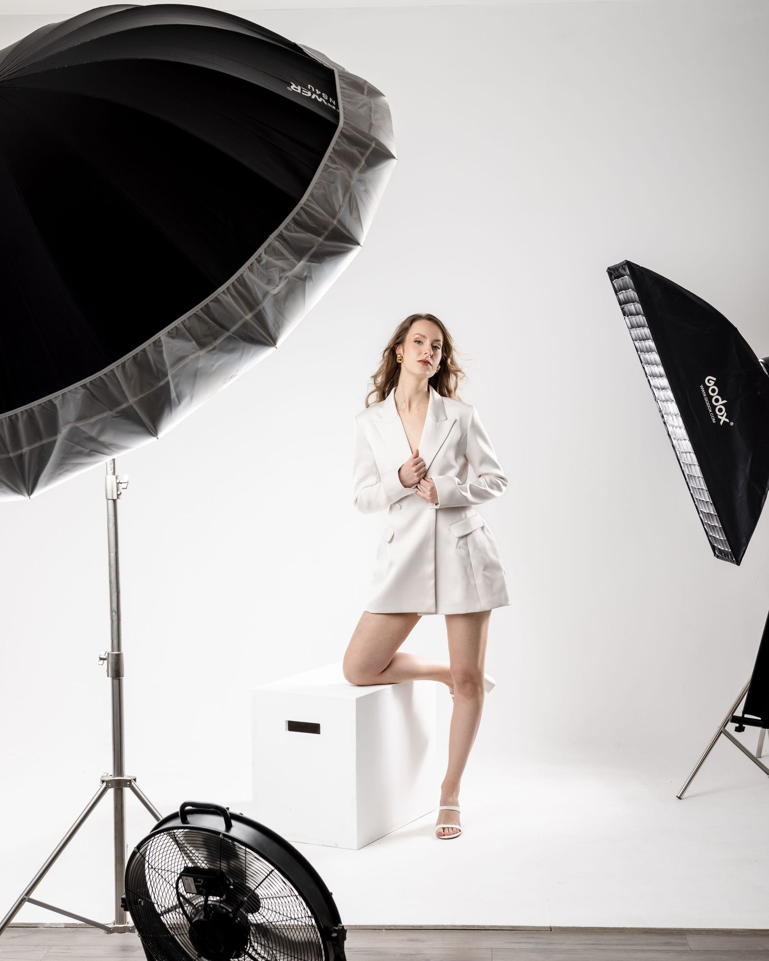 Woman in white blazer dress poses in photography studio with umbrella and softbox lights on white background.