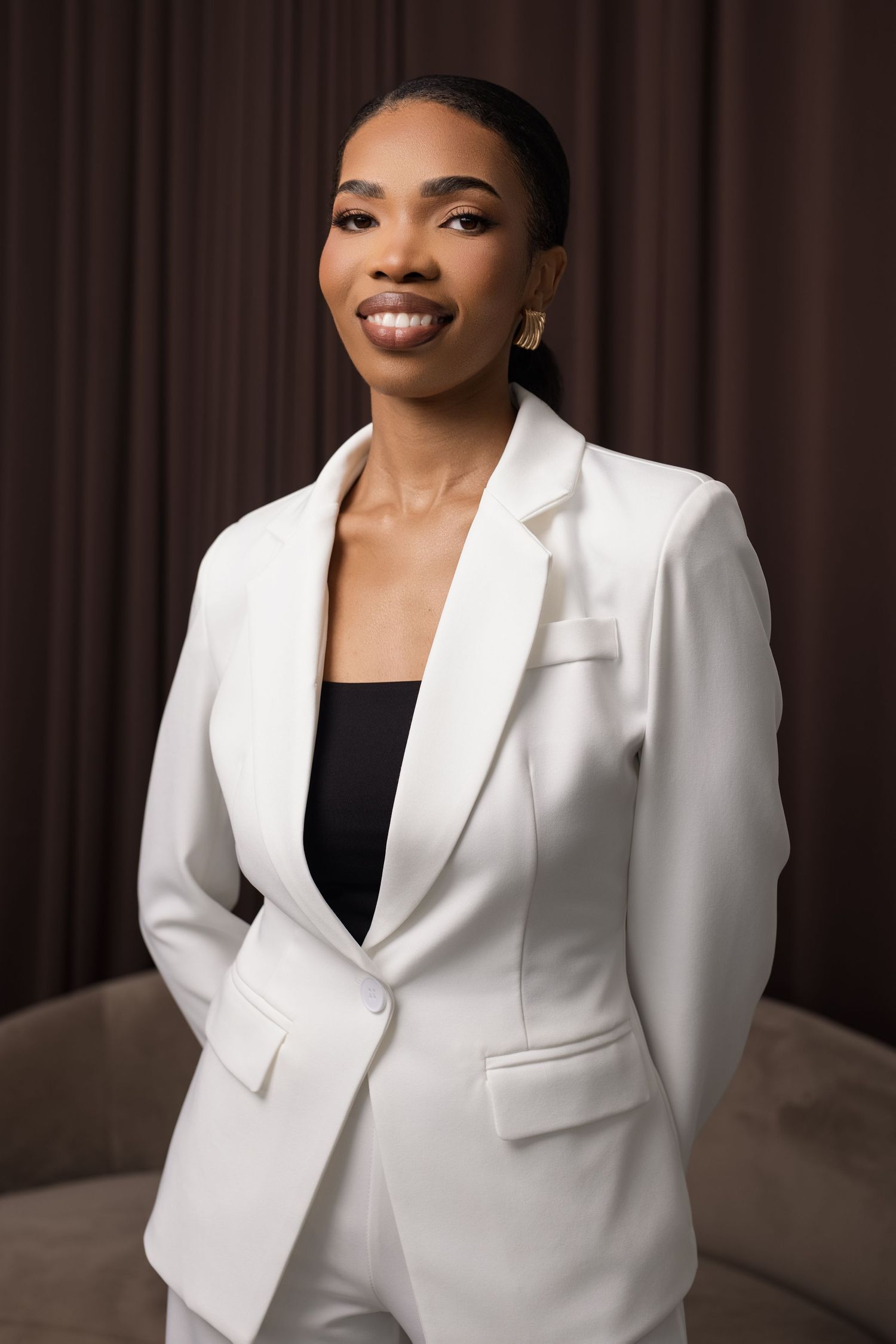 A confident young Black woman smiles in a white blazer suit and black top, posing against a brown curtain backdrop.