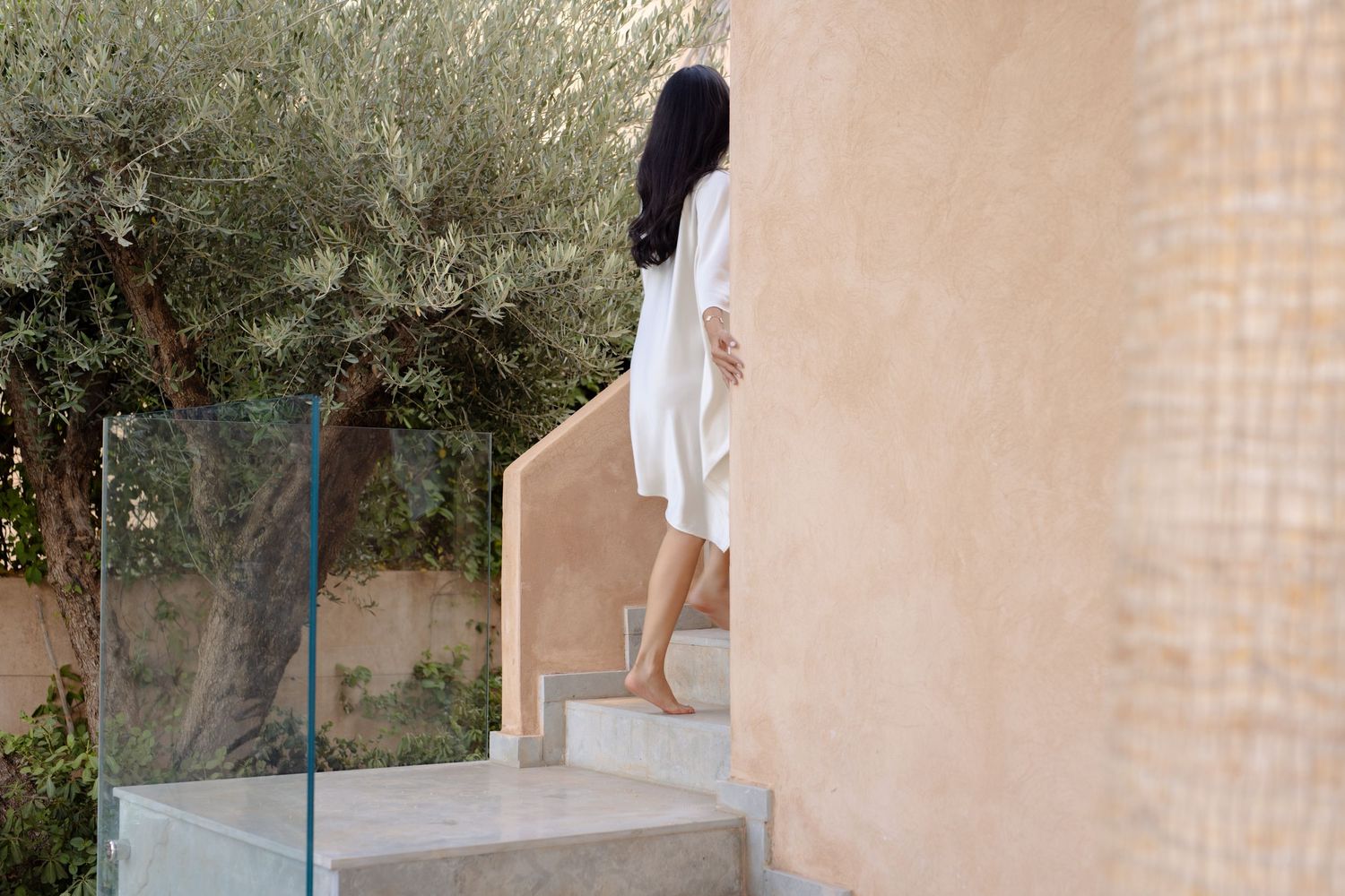 Woman in white dress walking barefoot up outdoor stone steps beside a glass railing and terracotta wall.
