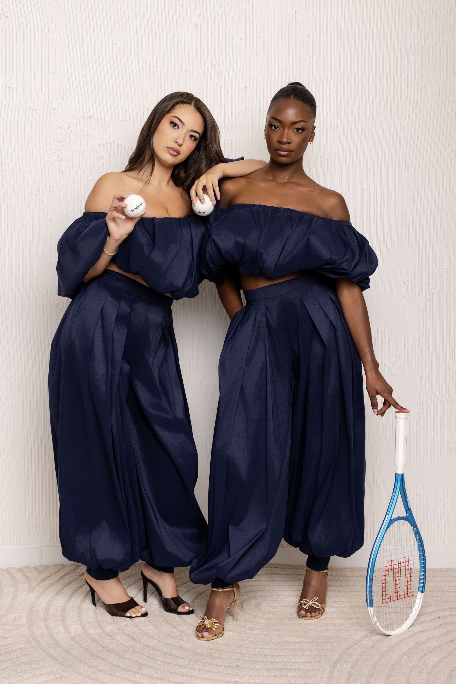 Two women in navy off-shoulder outfits pose with a tennis racket and ball against a white wall.