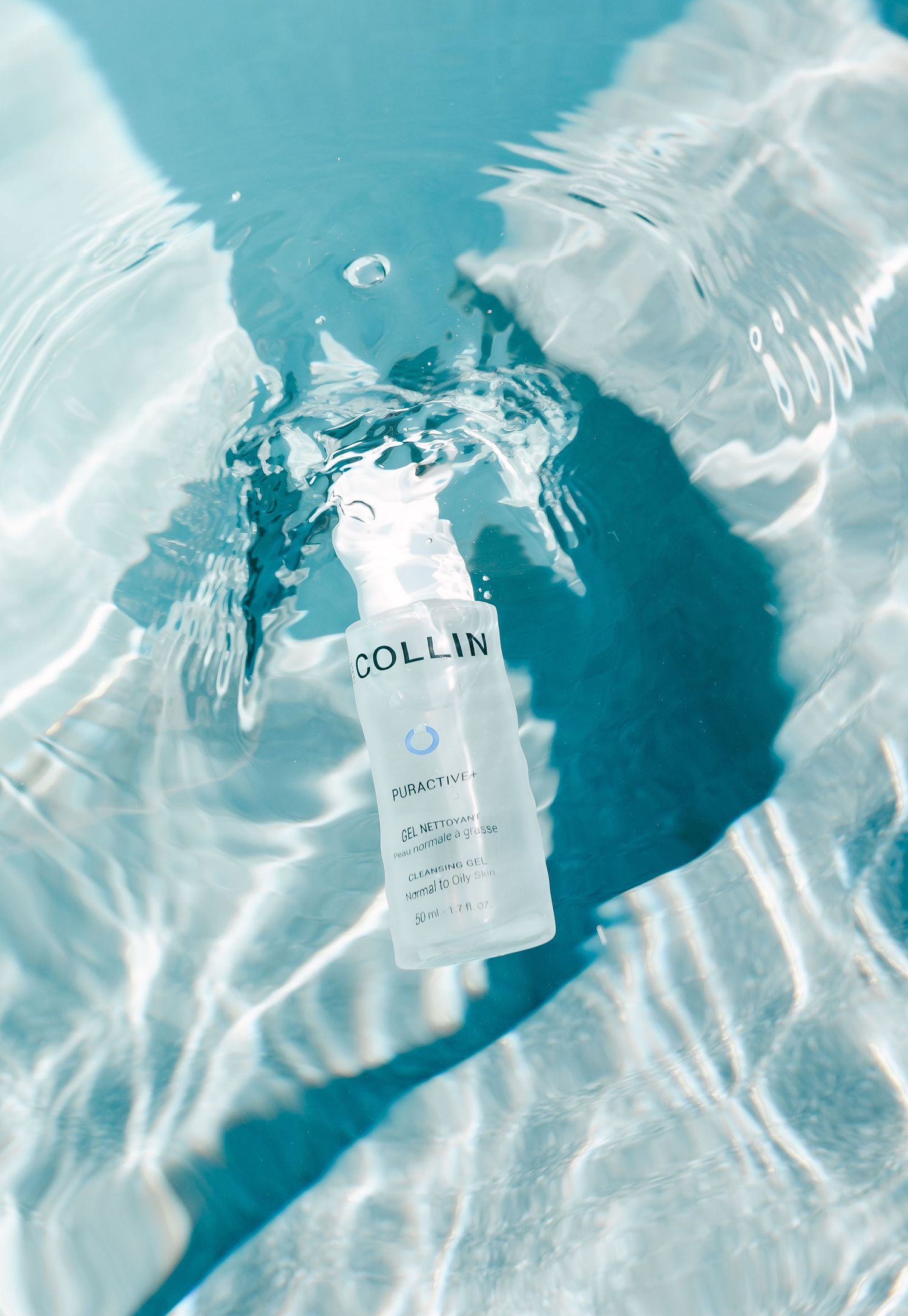 A bottle of G.M. COLLIN skincare product submerged in clear rippling water with turquoise blue tones.