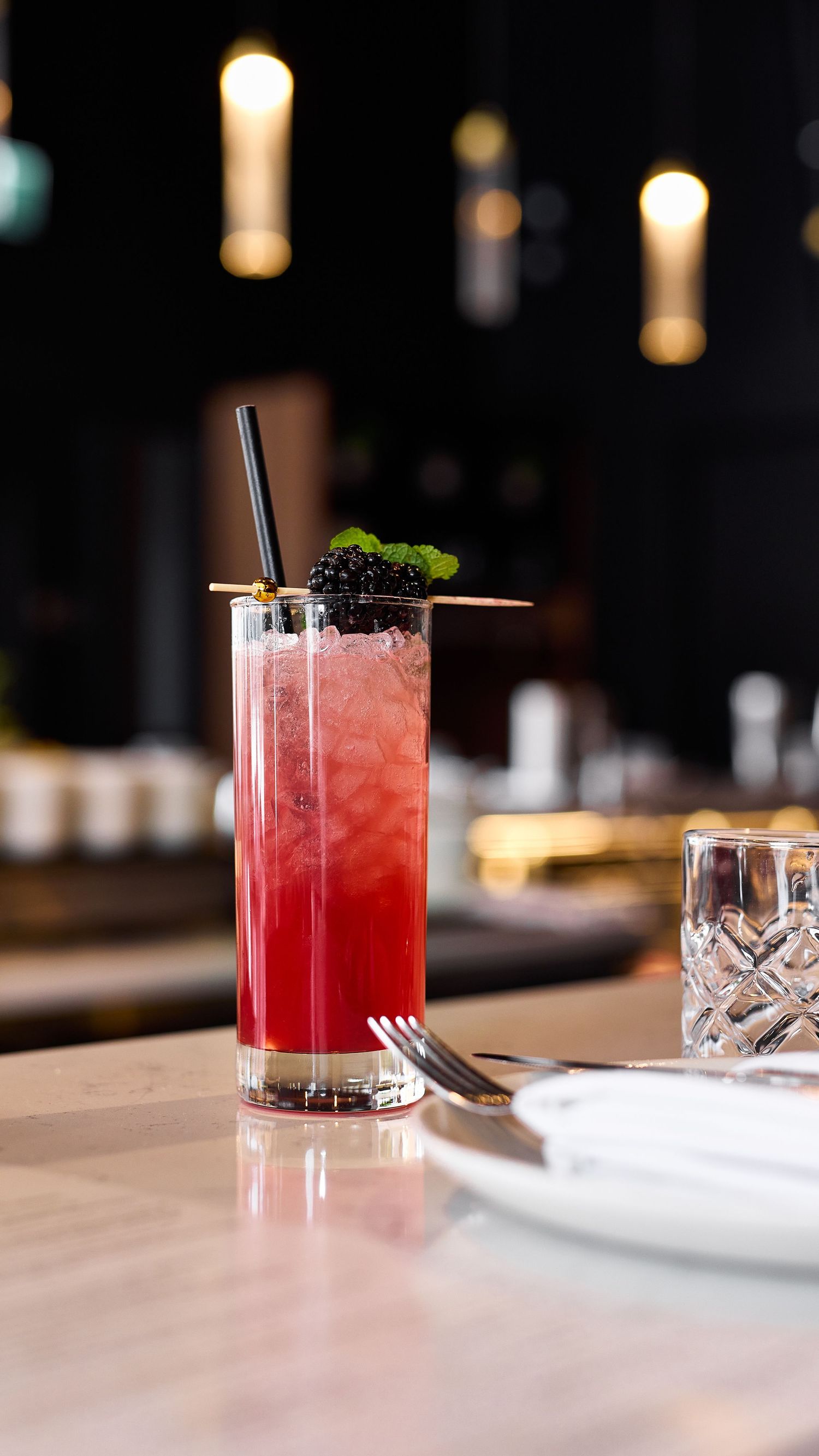 A vibrant red cocktail garnished with mint sits in a tall glass on a restaurant table with ambient lighting.