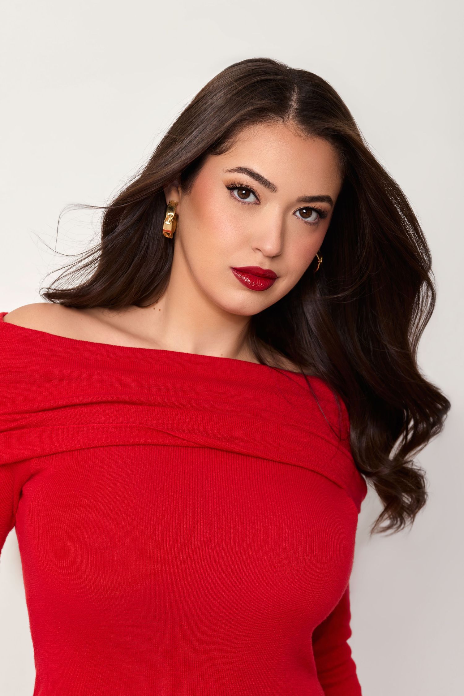 Young woman in red off-shoulder top with dark wavy hair and red lips poses against light background.