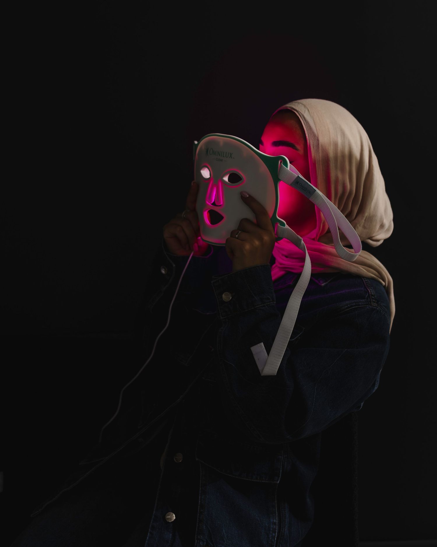 Person in a winter coat and head covering illuminated by dramatic magenta lighting against a dark background.