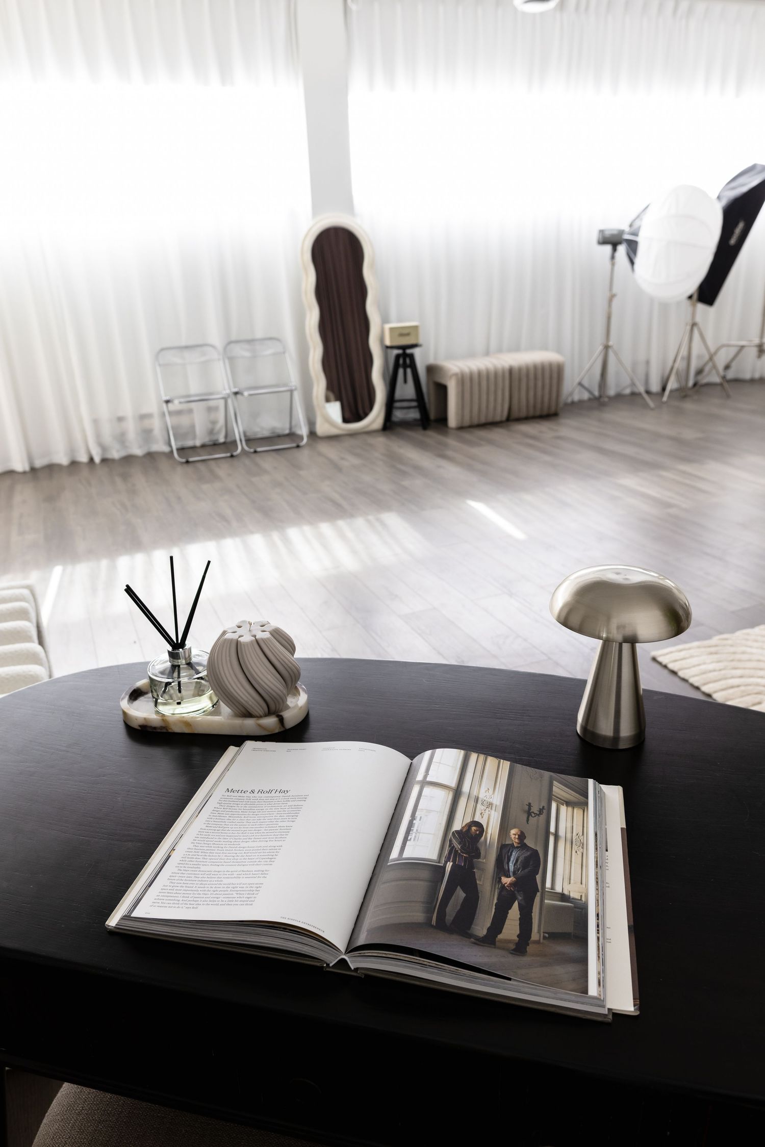 Modern photography studio with open magazine, reed diffuser, and mushroom lamp on dark table, soft lighting and props in background.
