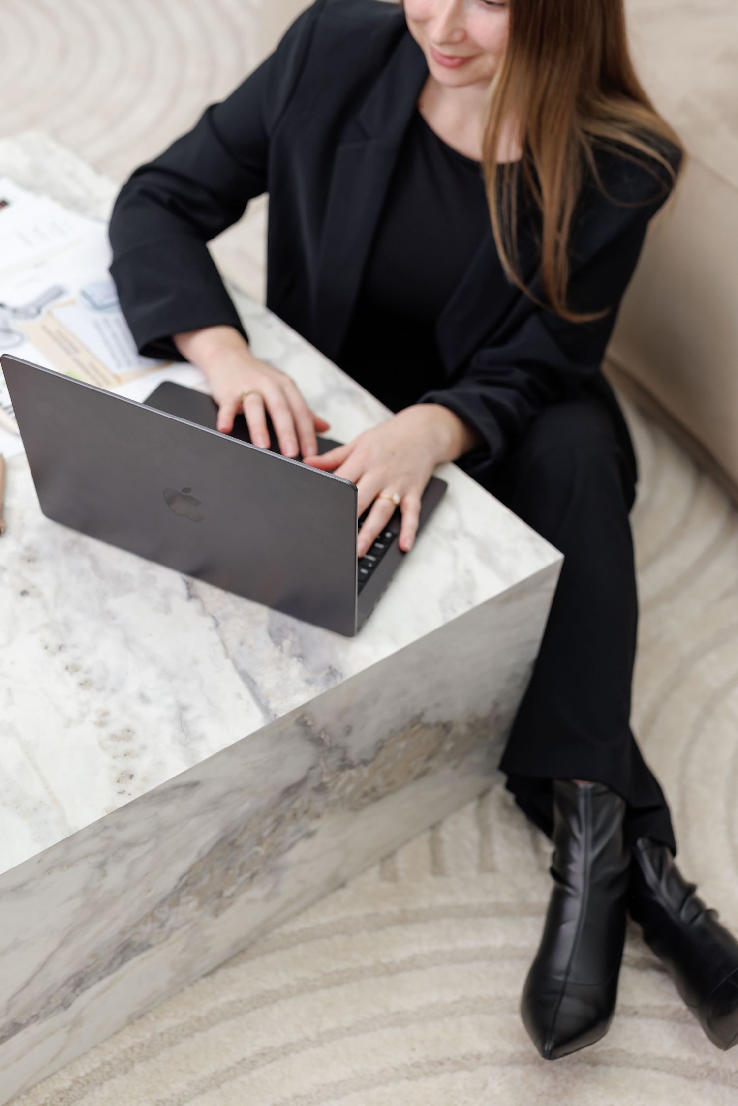 Professional woman in black suit working on laptop at marble table, wearing black boots in modern office setting.
