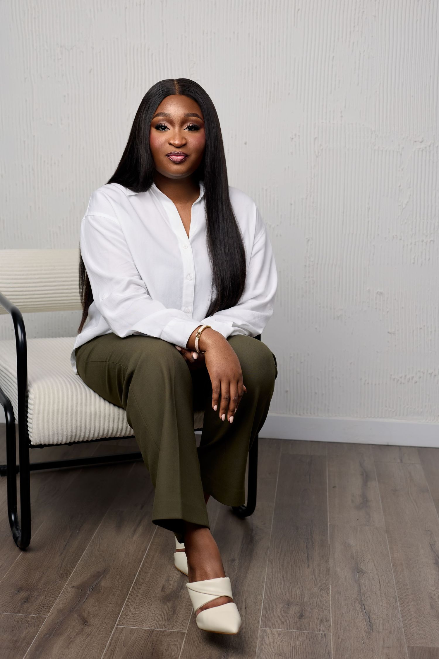 A person in a white button-down shirt and olive pants sits casually on a wooden floor against a light gray wall.