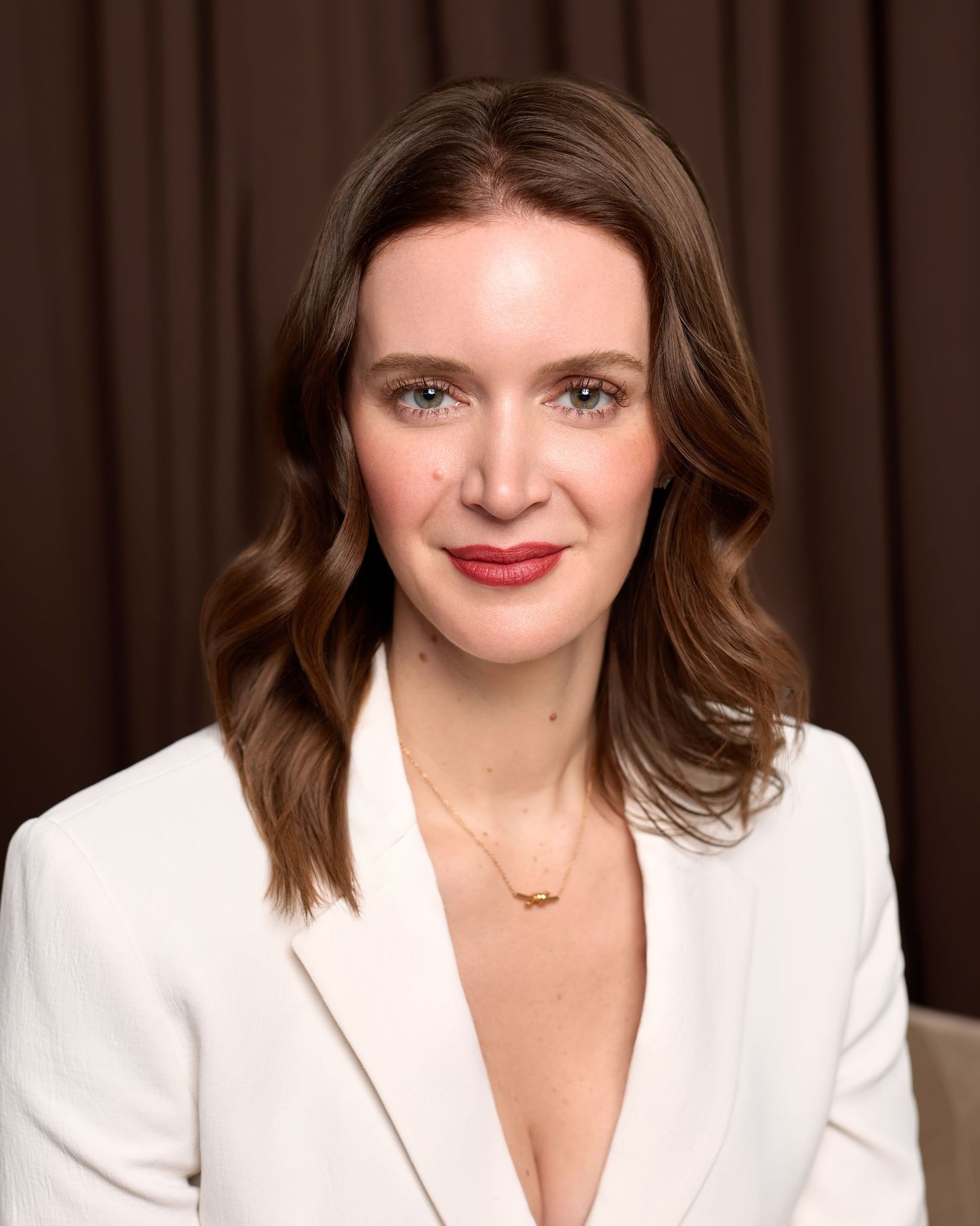 A woman with brown wavy hair and blue eyes wearing a white blazer and gold necklace poses for a portrait.
