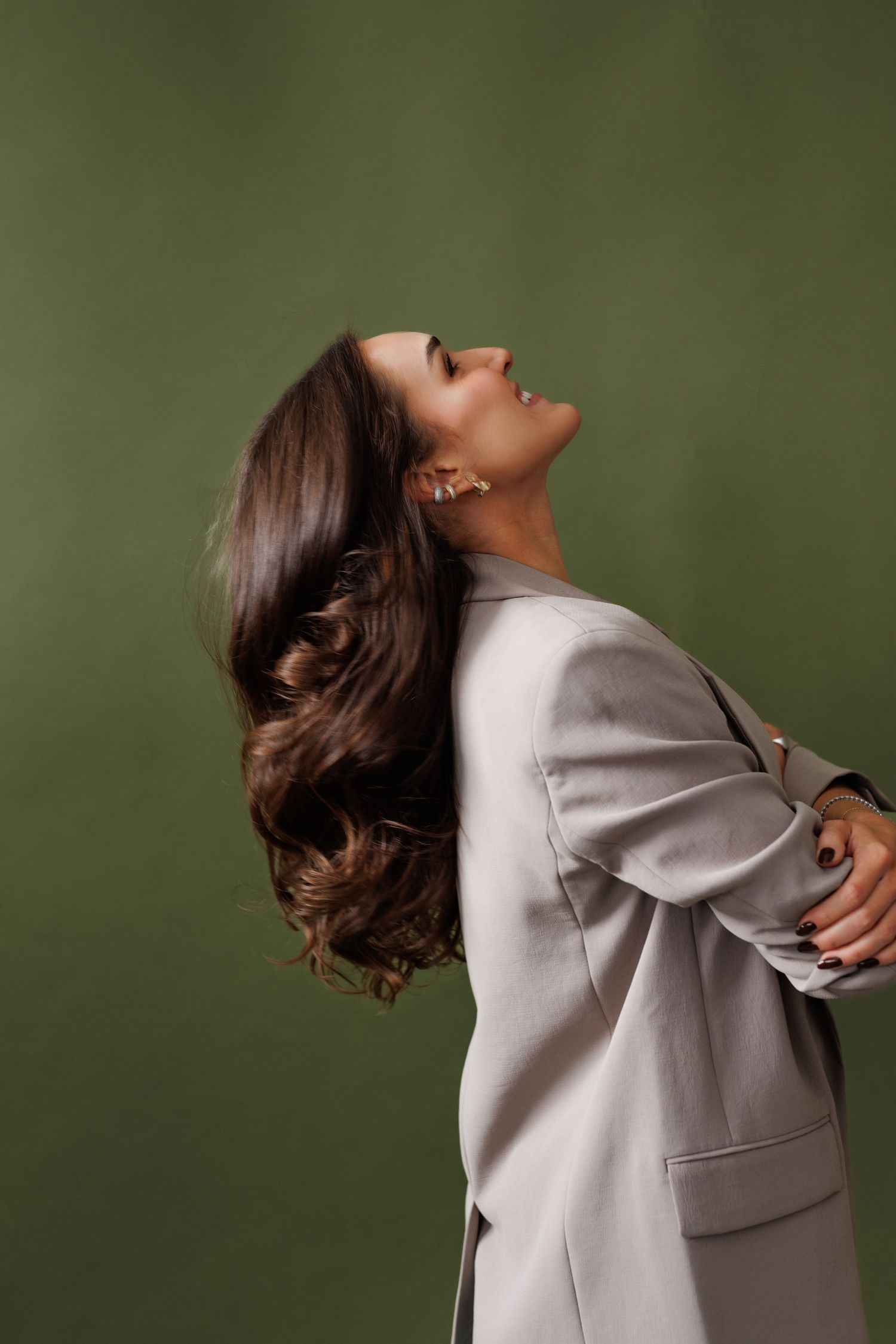 A woman in a light gray blazer poses with flowing brunette hair against an olive green background.