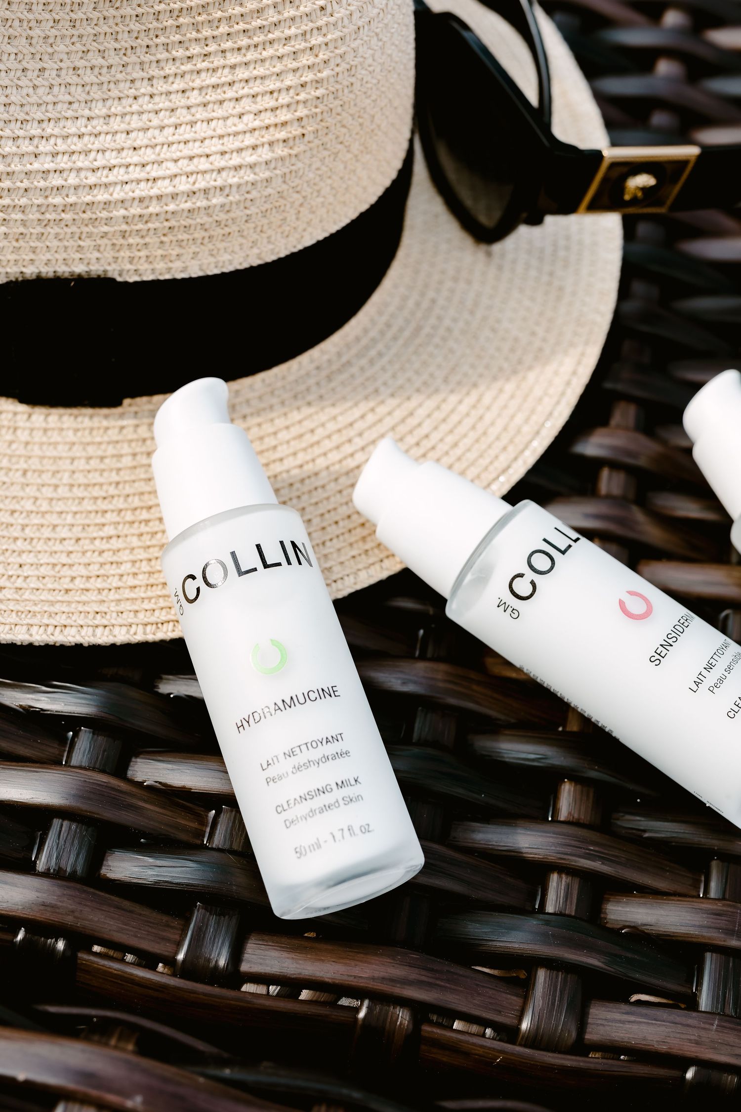 Two Collin skincare bottles displayed on a woven surface with a straw hat featuring a black band.