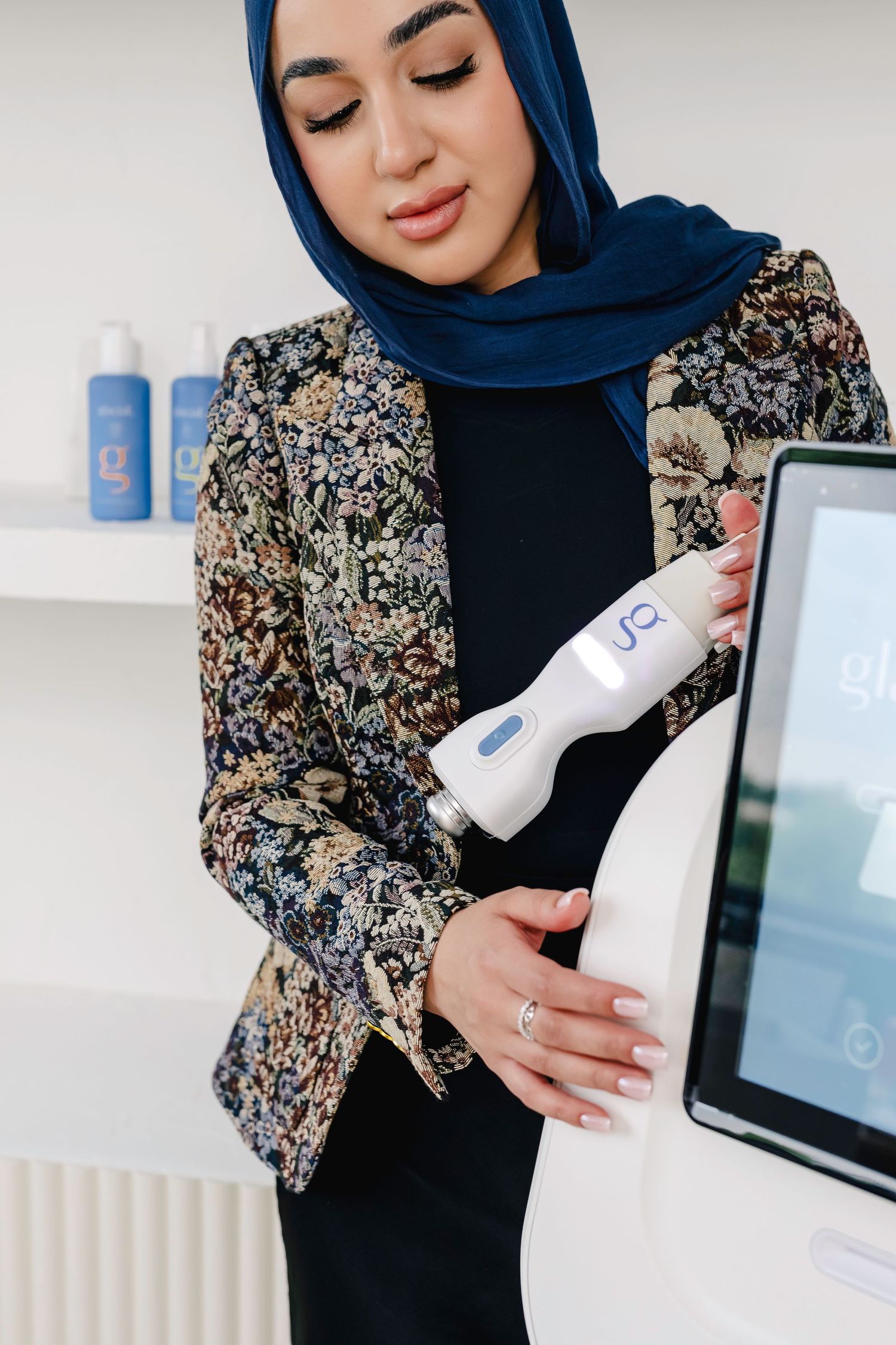 Person wearing navy hijab and floral print jacket holds skincare product while standing near laptop display.