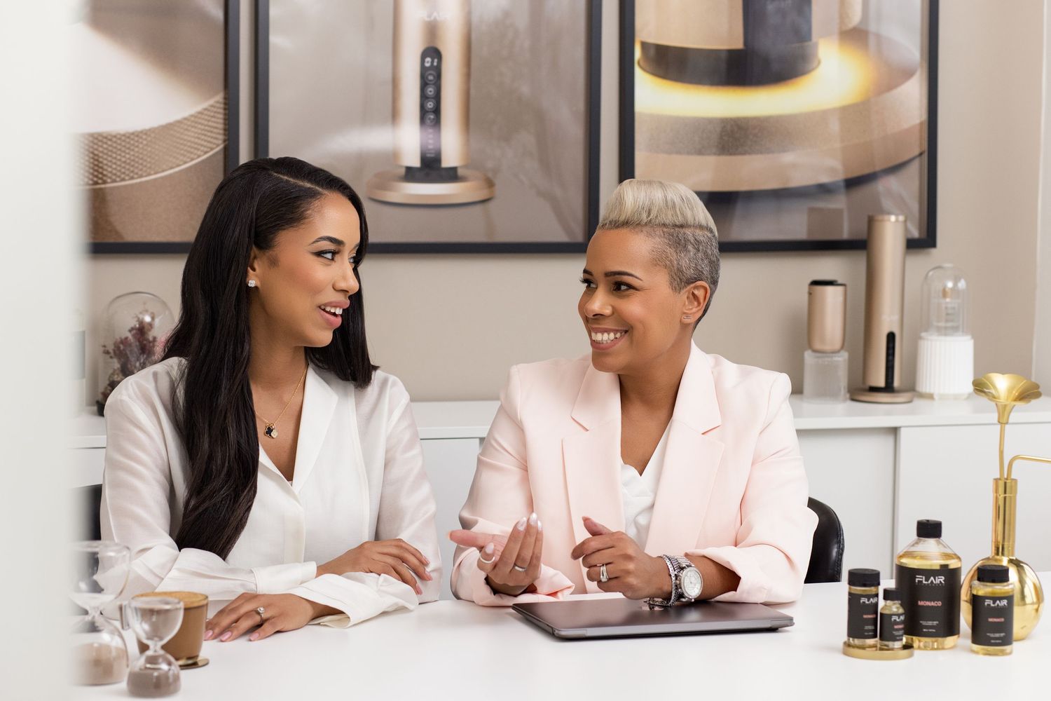 Two women in elegant attire smile at a white table with luxury beauty products in a modern studio.