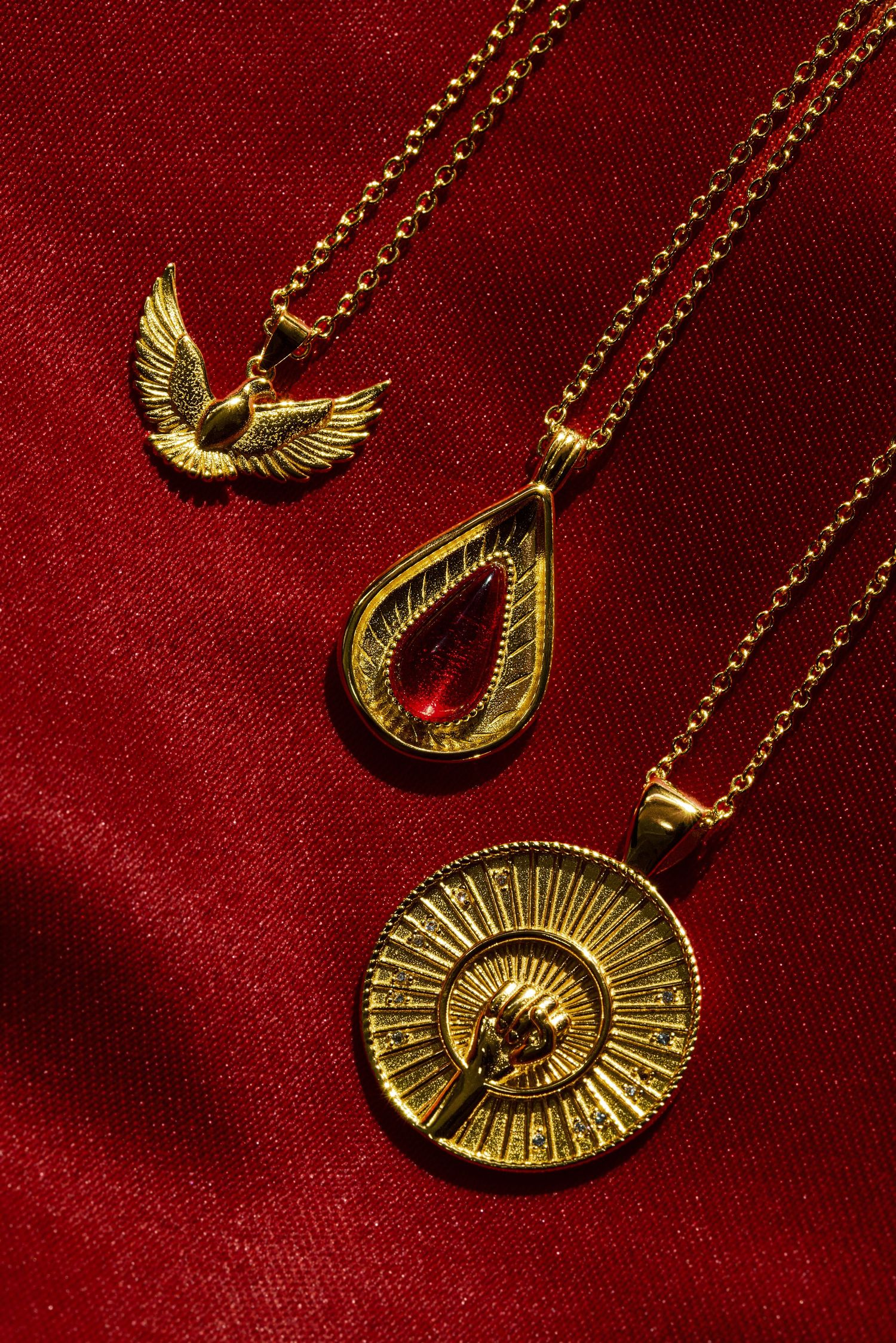 Three gold necklaces on red satin: winged bird pendant, teardrop with red gem, and circular sunburst medallion.