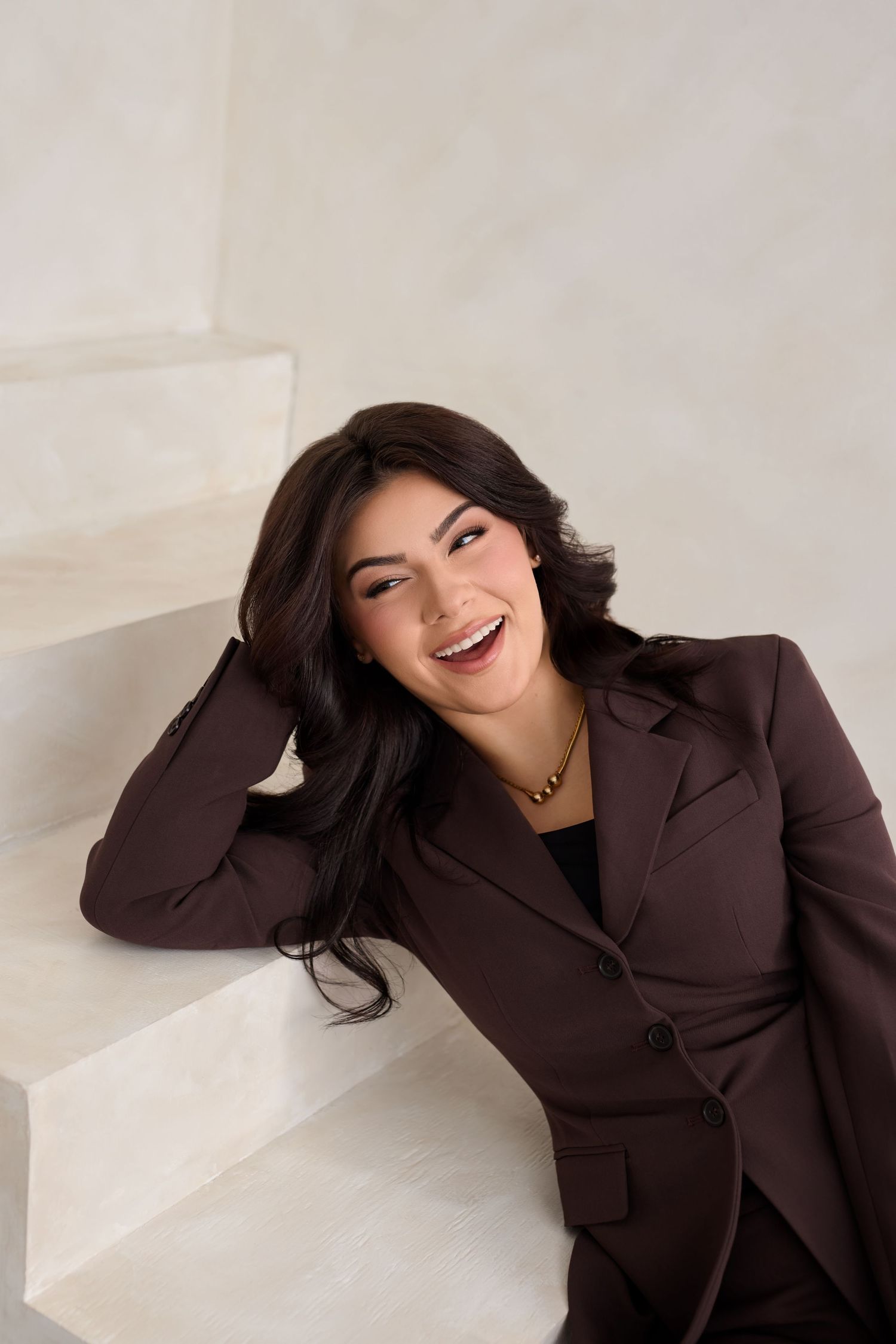 Professional portrait photo of someone in a brown blazer leaning against white stairs while smiling warmly.