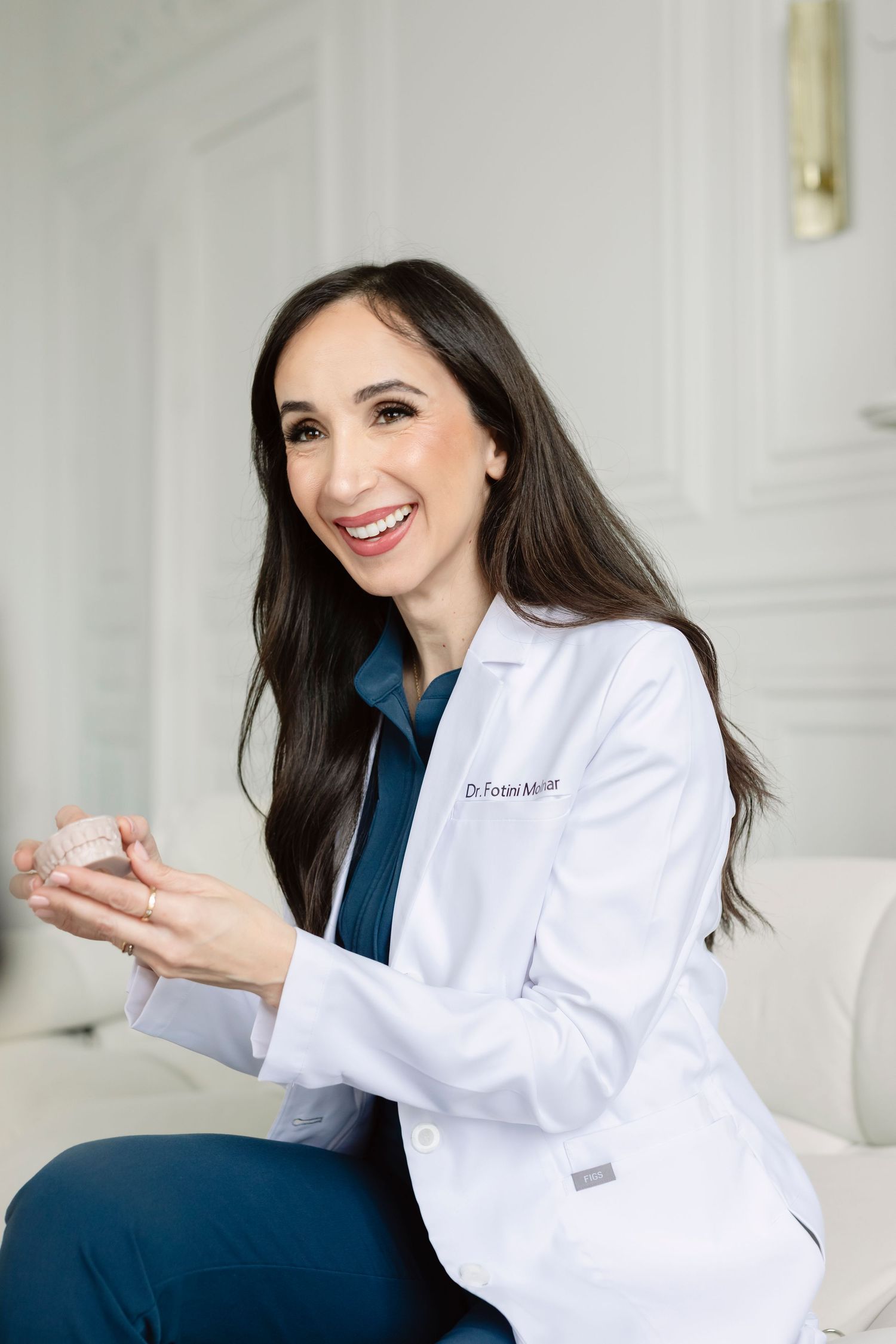 Smiling female doctor in white lab coat holding dental aligner model, seated in bright modern medical office.