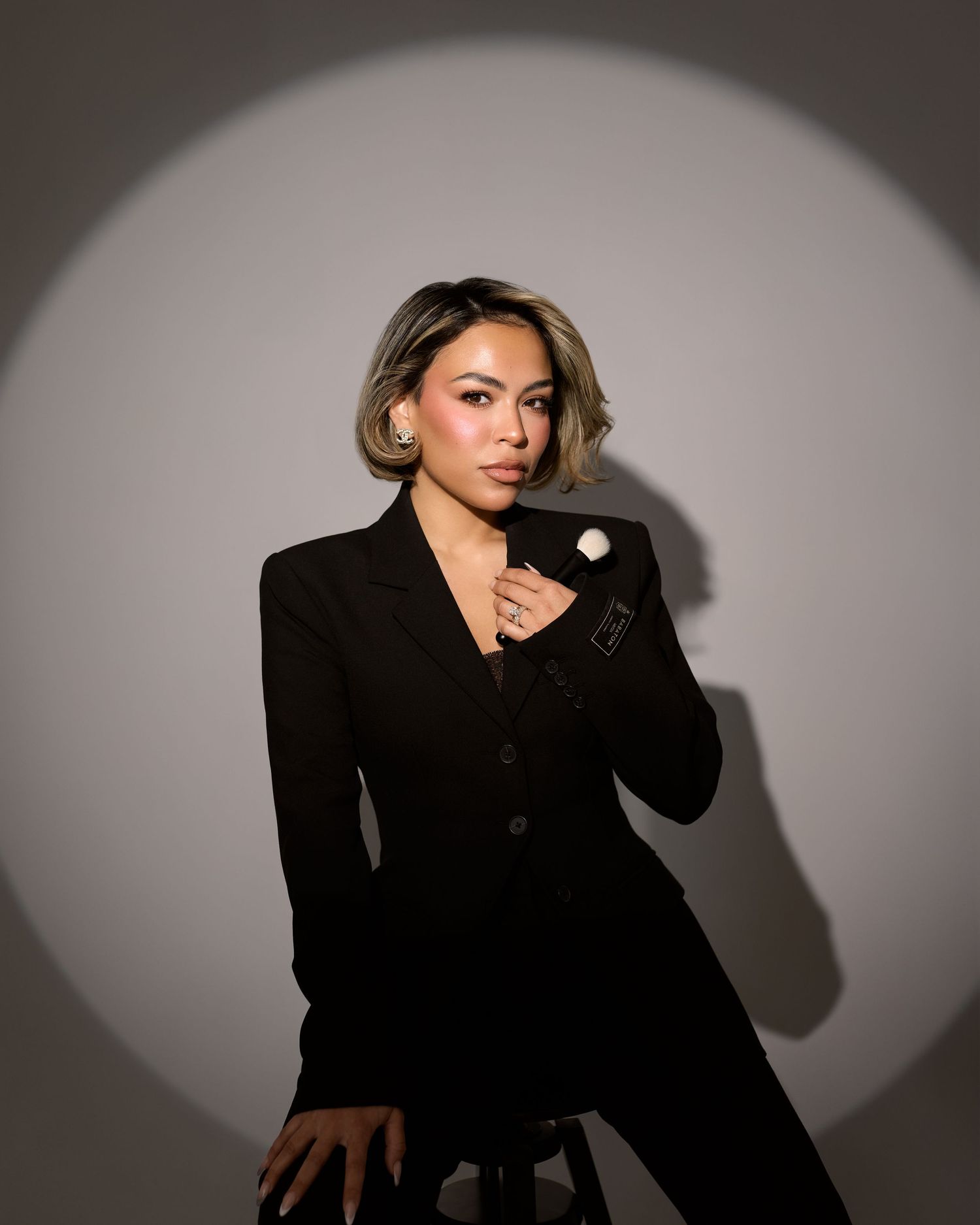 A model in a black blazer poses dramatically against a gray background with moody lighting and shadows.