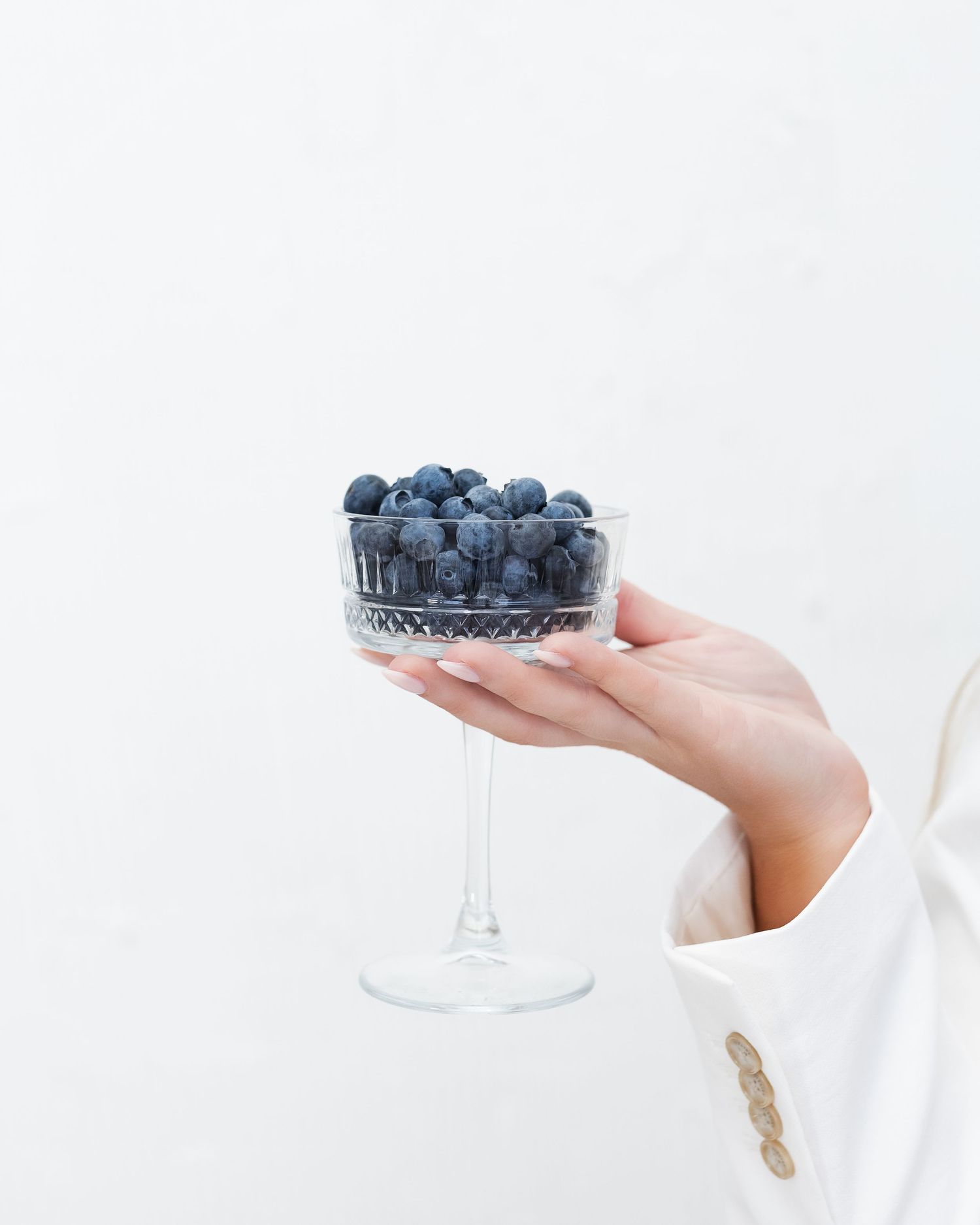 A hand holding a crystal coupe glass filled with fresh blueberries against a white background.