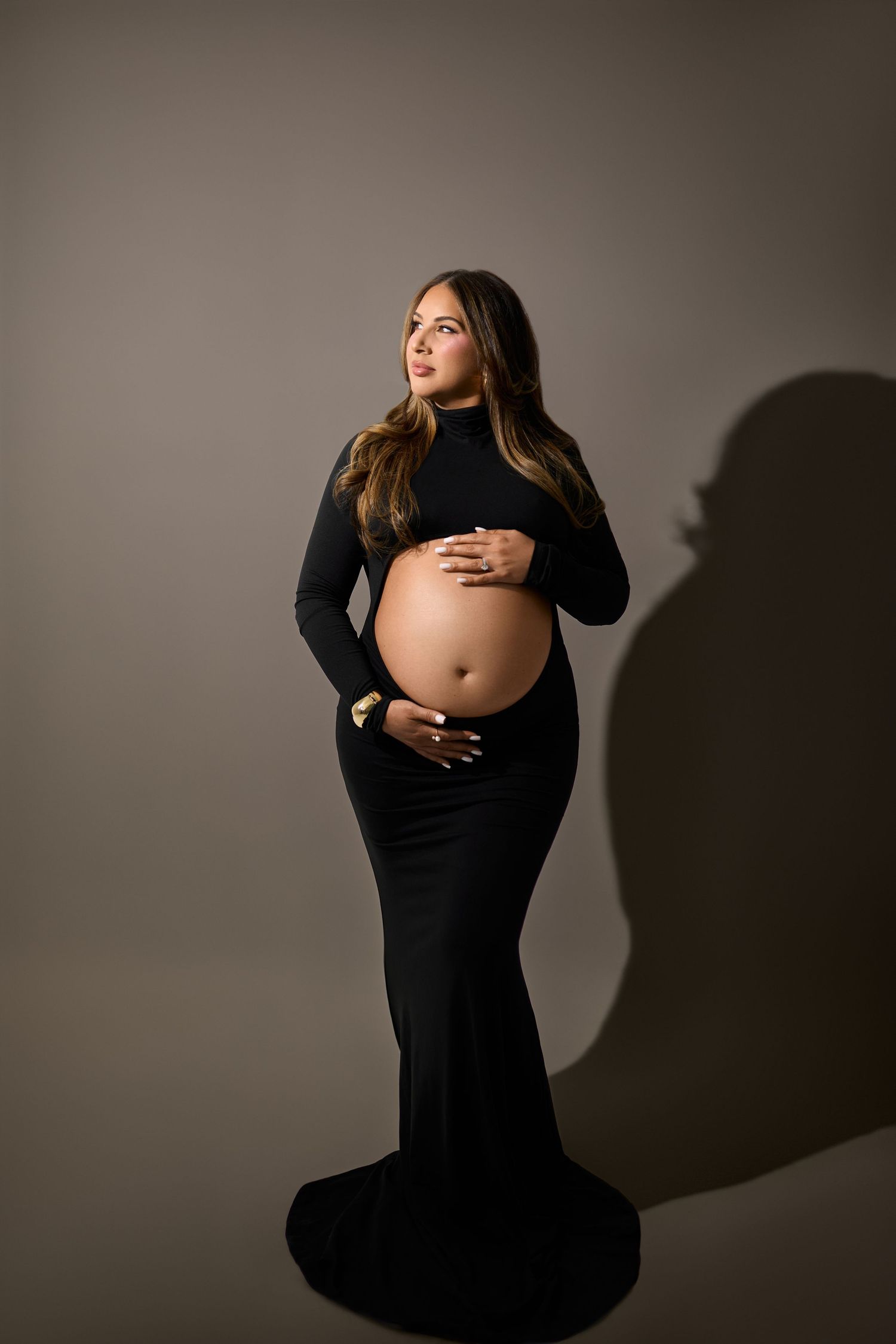A pregnant woman in a long black dress poses against a simple backdrop casting a dramatic shadow.