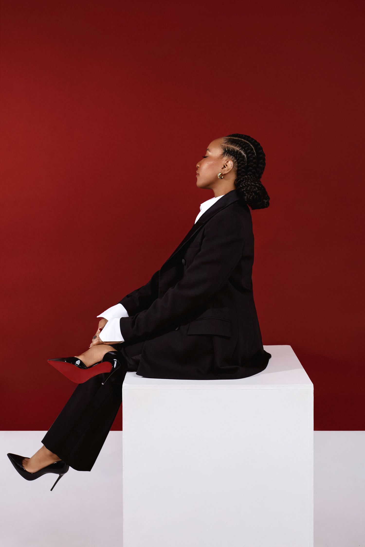 Figure in black blazer reclines on white geometric blocks against deep red backdrop creating minimalist fashion composition.