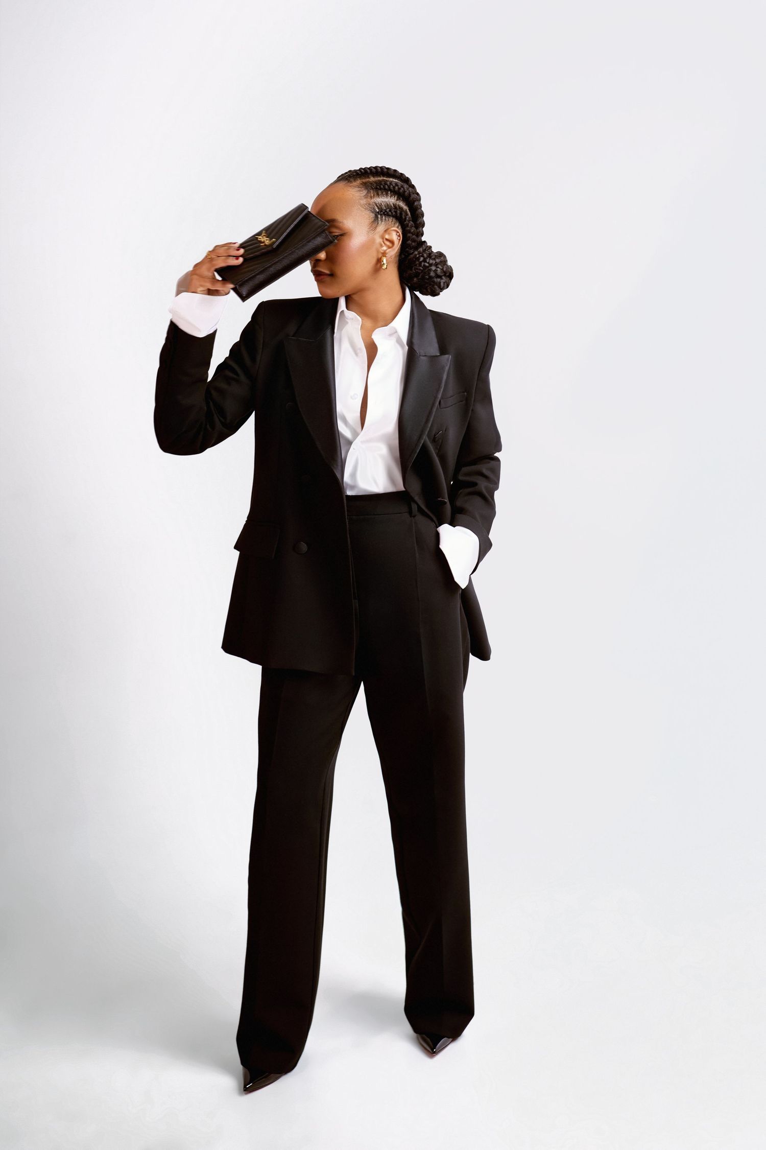 A woman in a black tailored suit poses confidently against a white background, holding a clutch purse near her face.