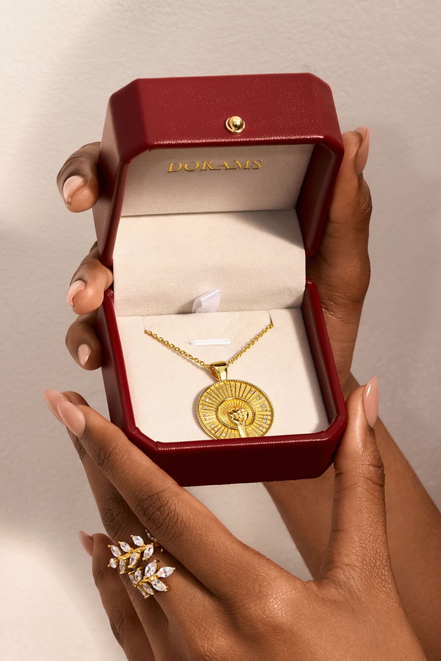 Hands holding and displaying an elegant gold pendant necklace in a burgundy velvet jewelry box.
