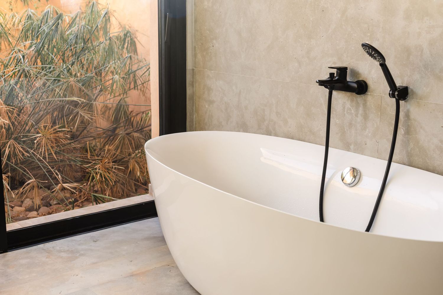 Modern freestanding cream bathtub with matte black faucet beside a framed botanical art panel in a minimalist bathroom.