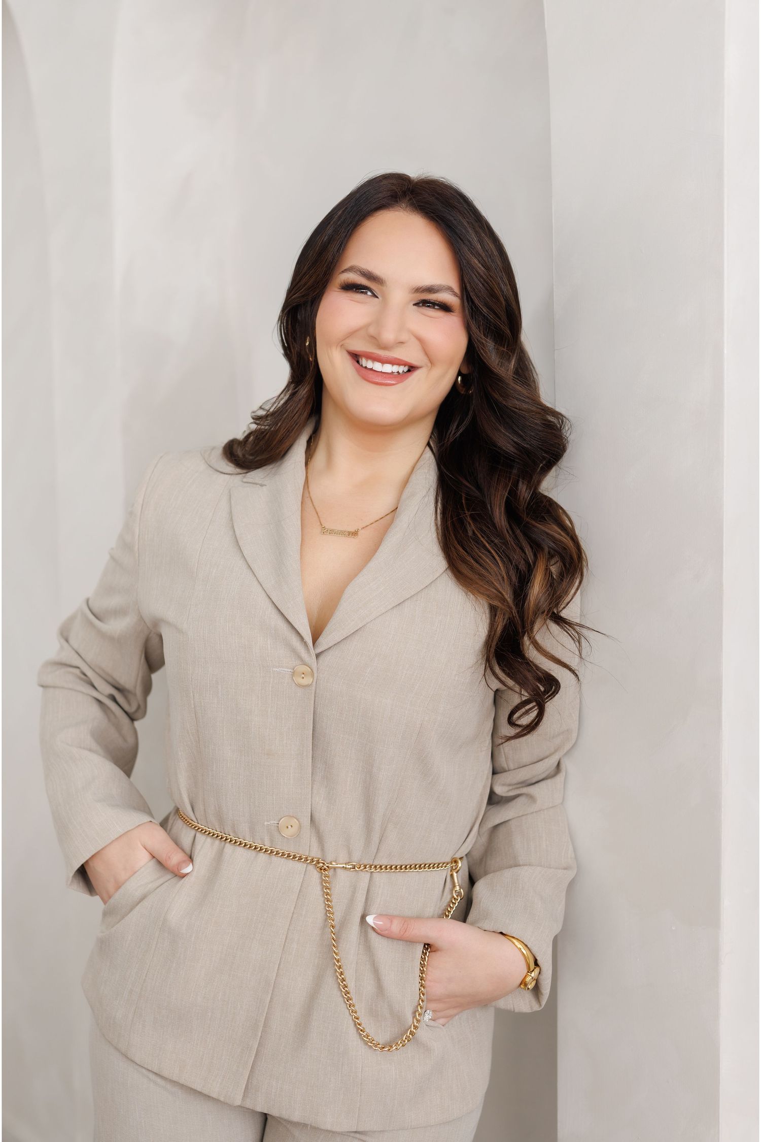 Professional studio portrait of someone wearing a beige blazer with a gold chain belt against a light gray background.