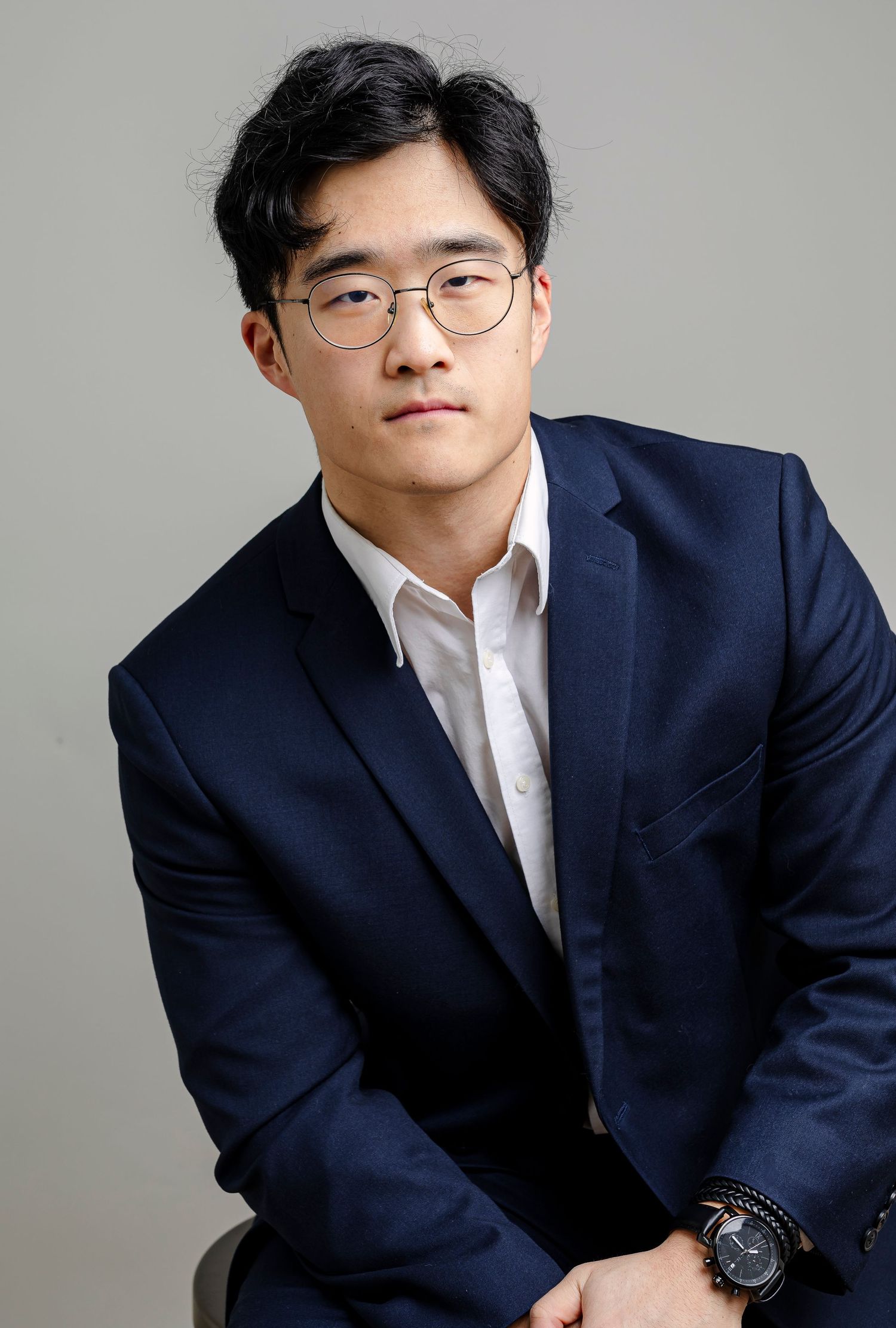 Professional headshot of someone wearing a navy suit and glasses against a light gray studio background.
