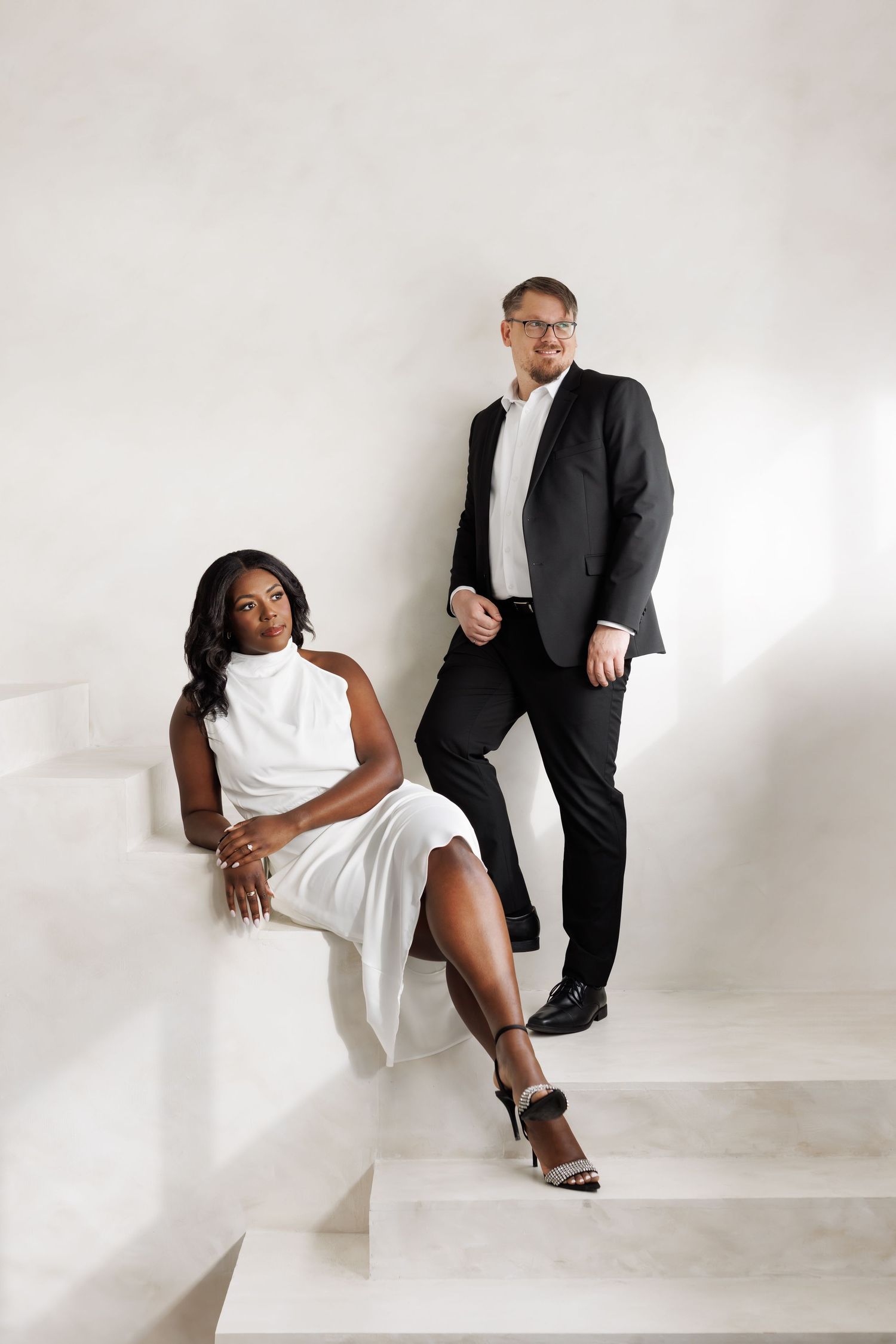 A couple in contrasting black and white outfits pose elegantly on white stairs during a minimalist photoshoot.