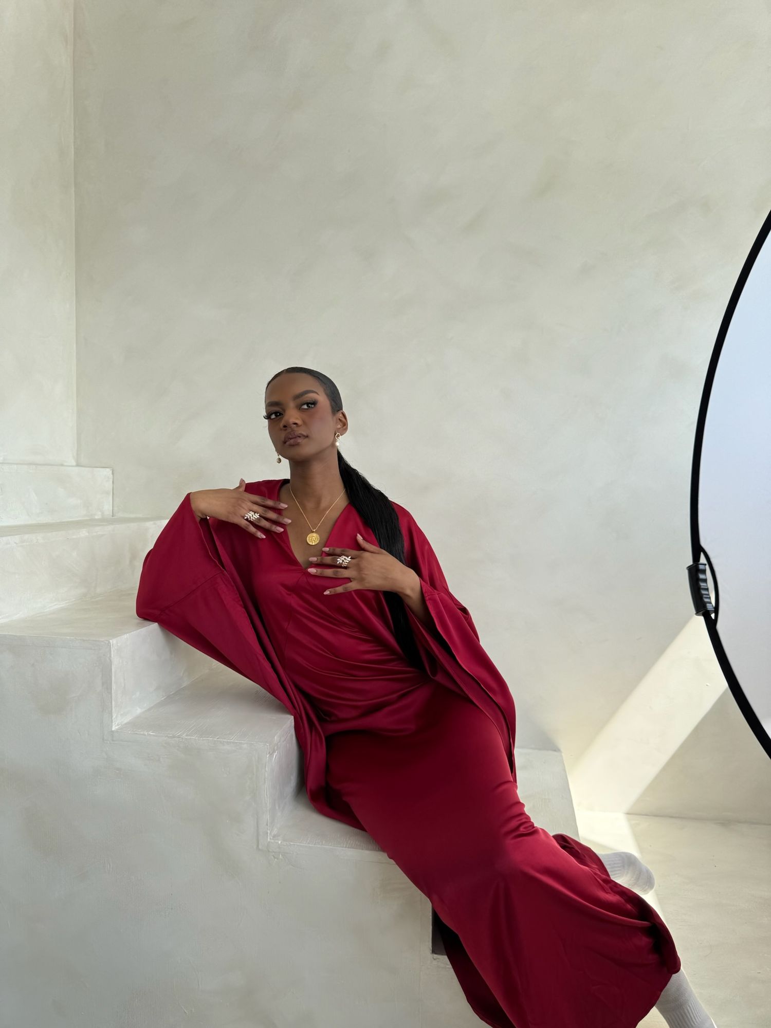 Model in burgundy dress poses dramatically against white studio wall.