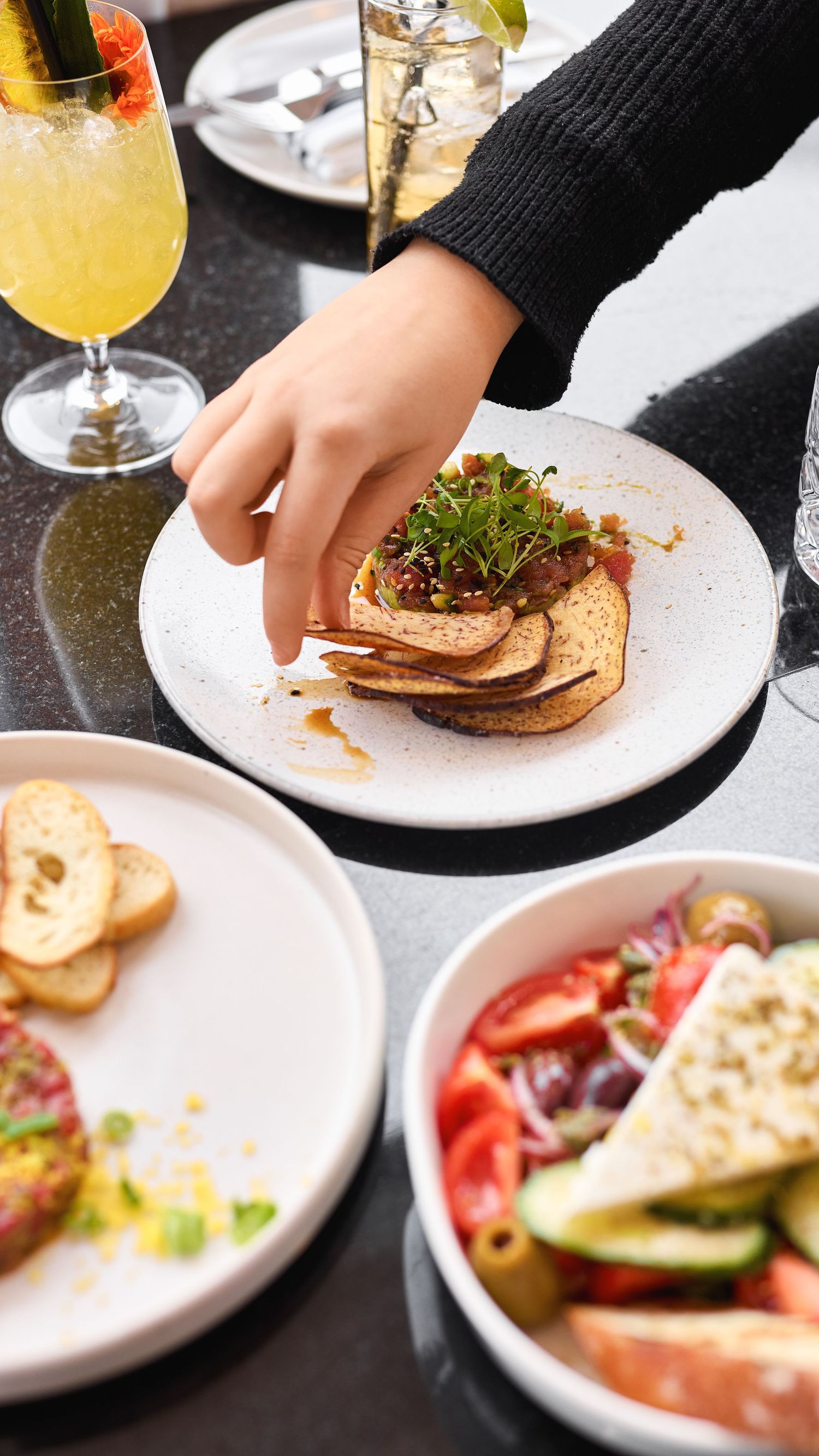 A hand reaches for appetizers on white plates with cocktails, featuring toasted bread, salad, and colorful drinks.