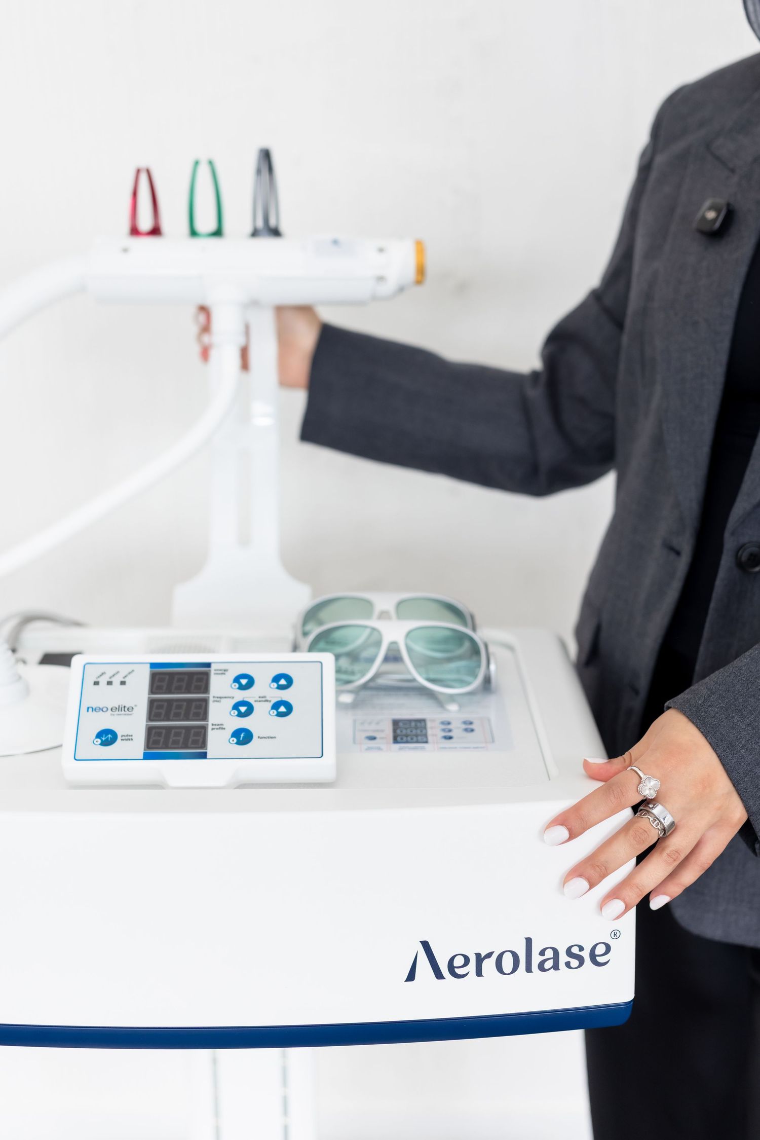 Woman in gray blazer operating an Aerolase laser device with protective eyewear and colorful handpieces in a clinic.