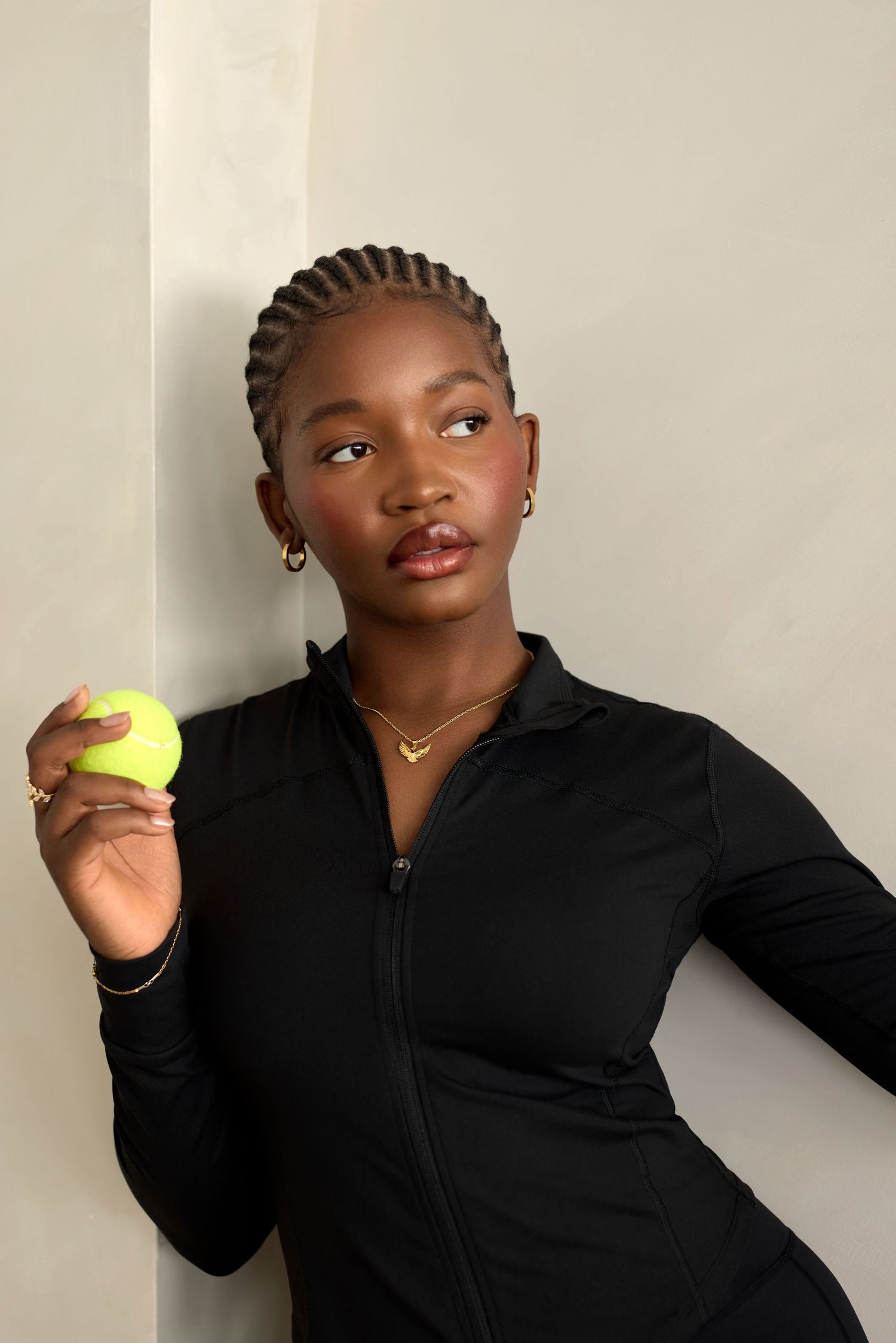 Person in black jacket holds yellow tennis ball while posing against light colored wall.