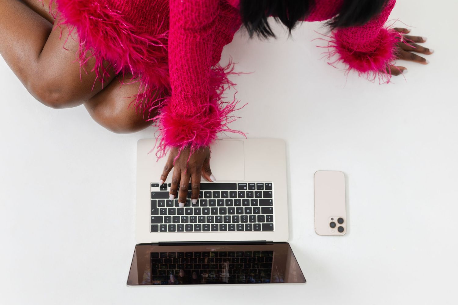 Person typing on laptop keyboard with bright pink feather boa draped over hands creating playful workspace atmosphere.