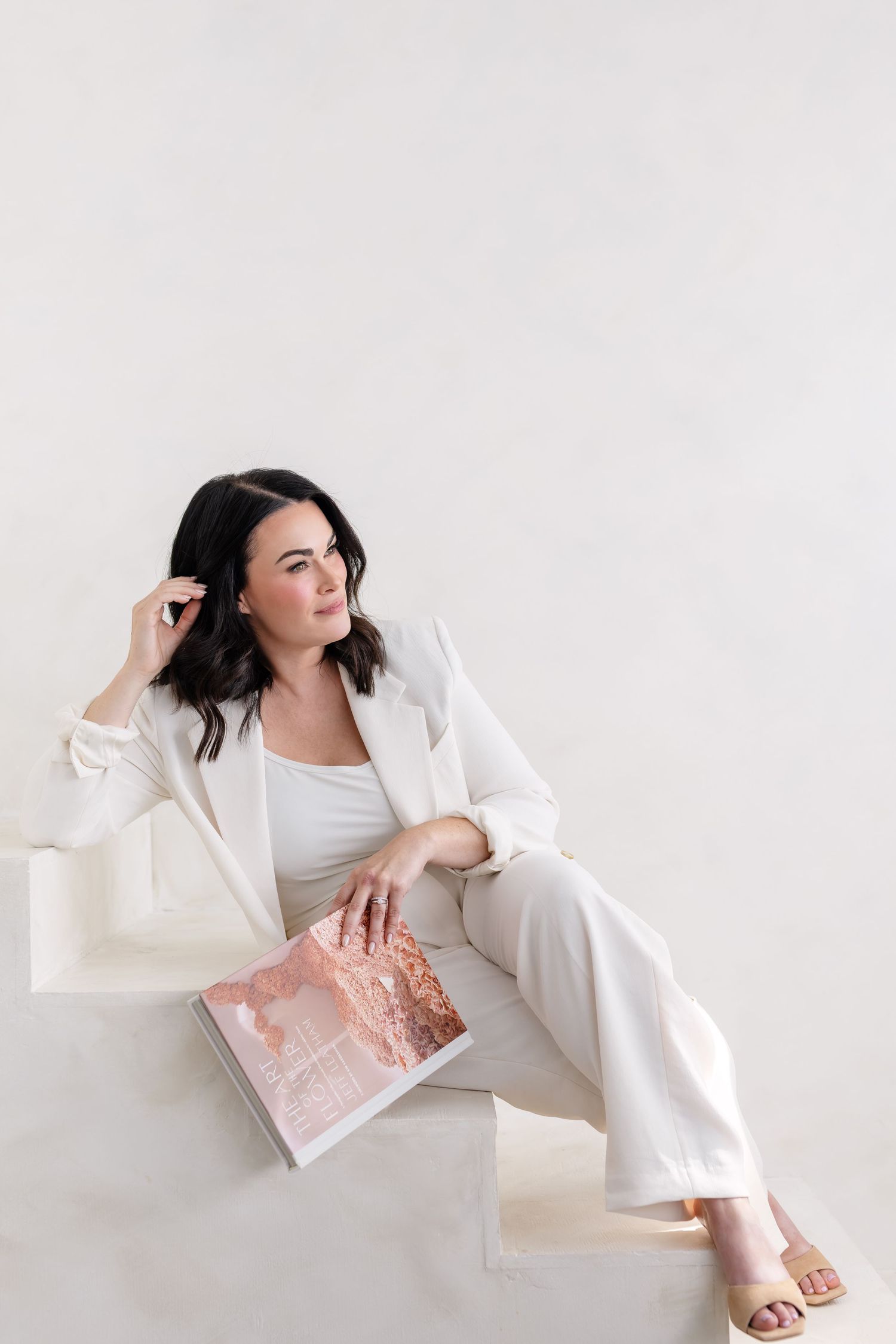 Woman in white suit lounges elegantly against white background while reading a pink book in artistic poses.