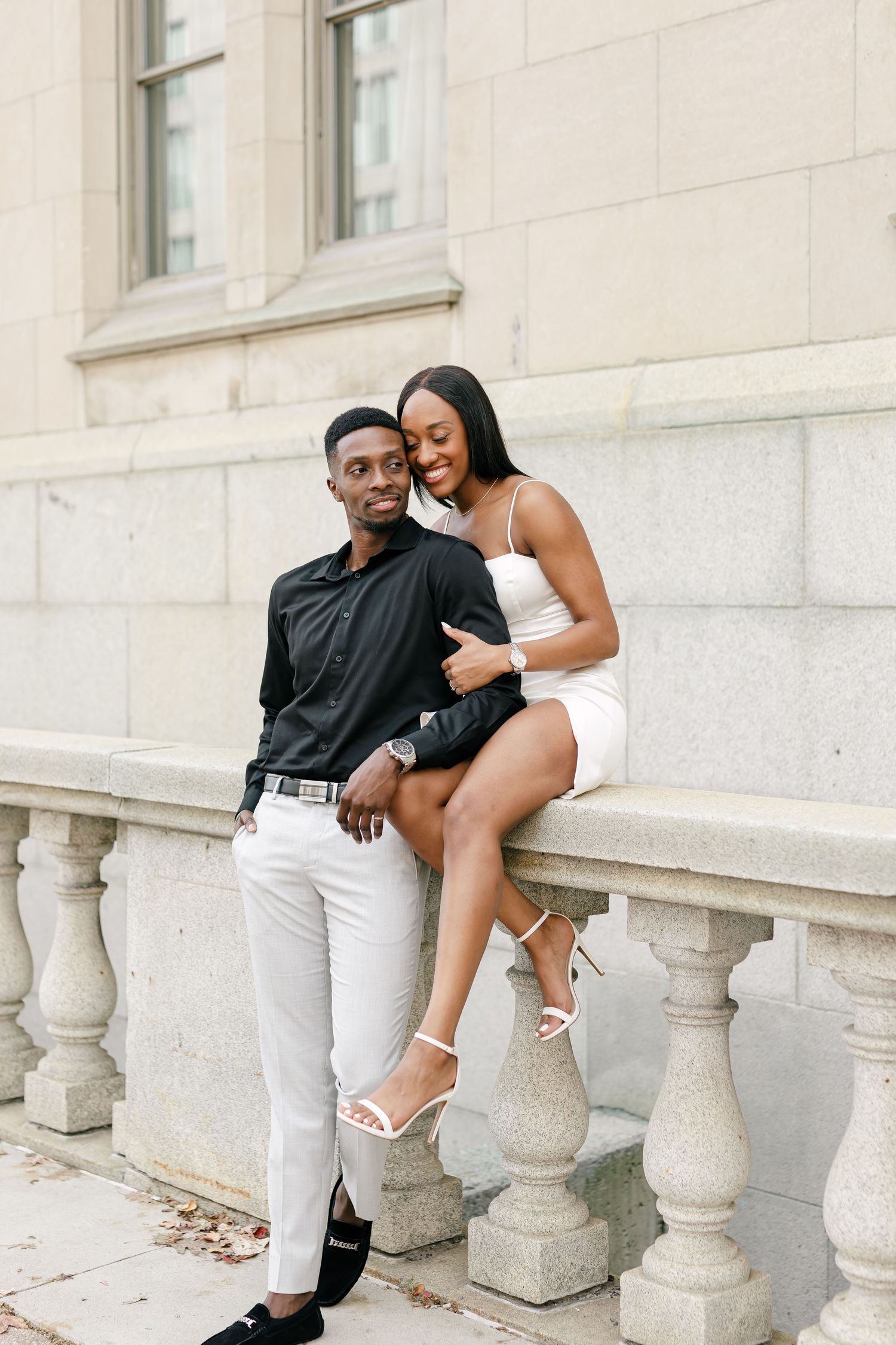 Couple shares romantic moments on stone balustrade in formal attire against white building exterior.