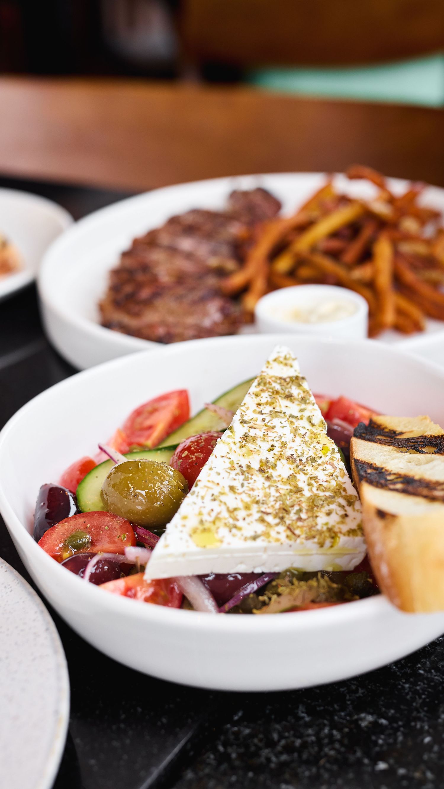Greek salad with feta cheese, olives, tomatoes, and pickles served alongside crispy french fries on a white plate.