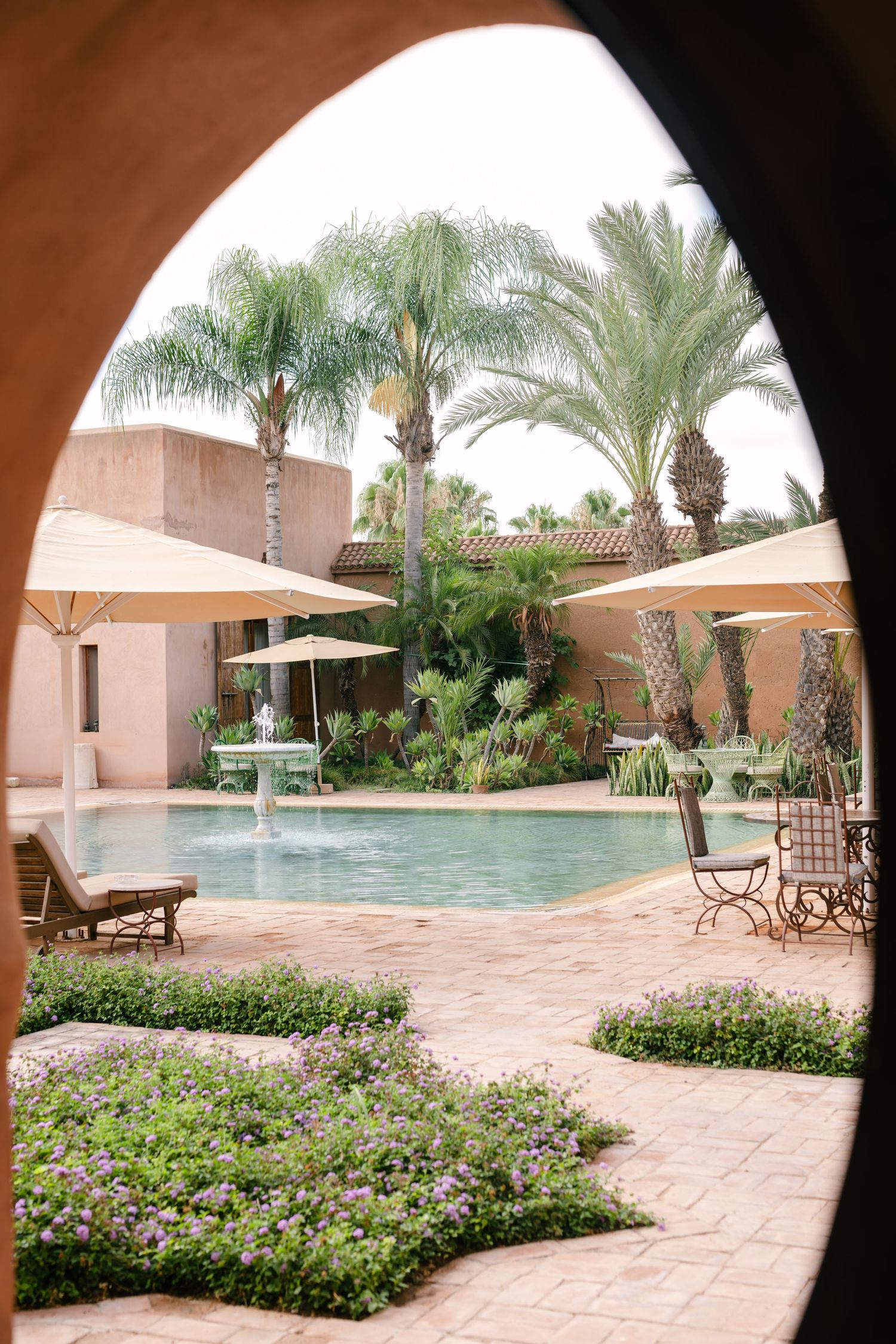 Luxury Moroccan resort pool with palm trees, umbrella shaded seating, and flowering gardens viewed through an arch.