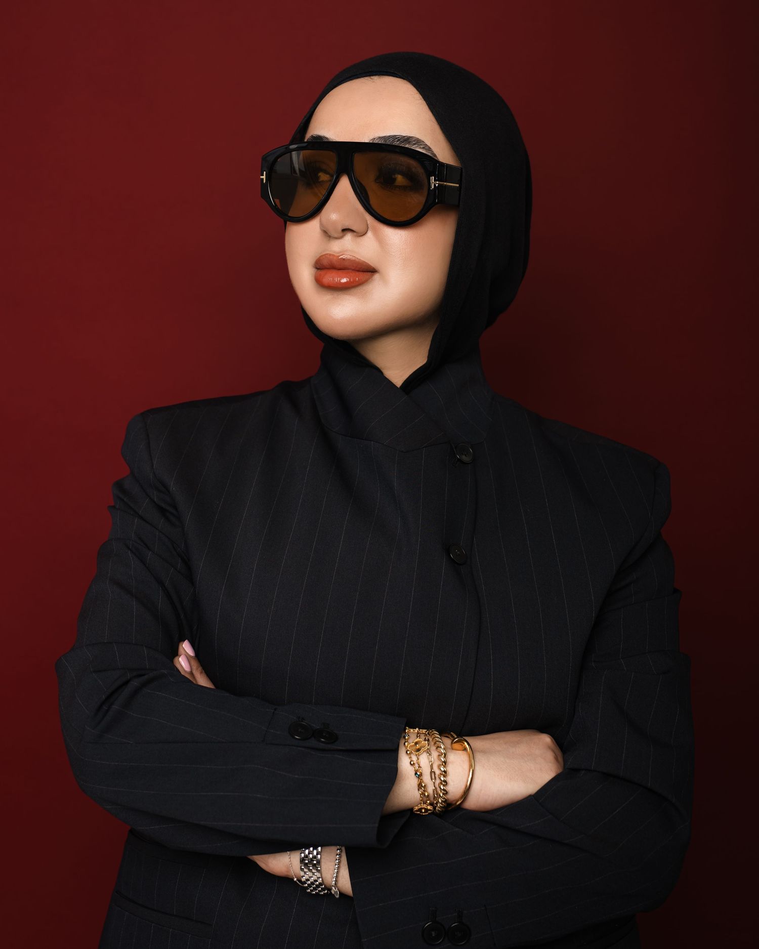 Woman in black hijab and blazer wearing oversized sunglasses and gold bracelets, arms crossed against dark red background.