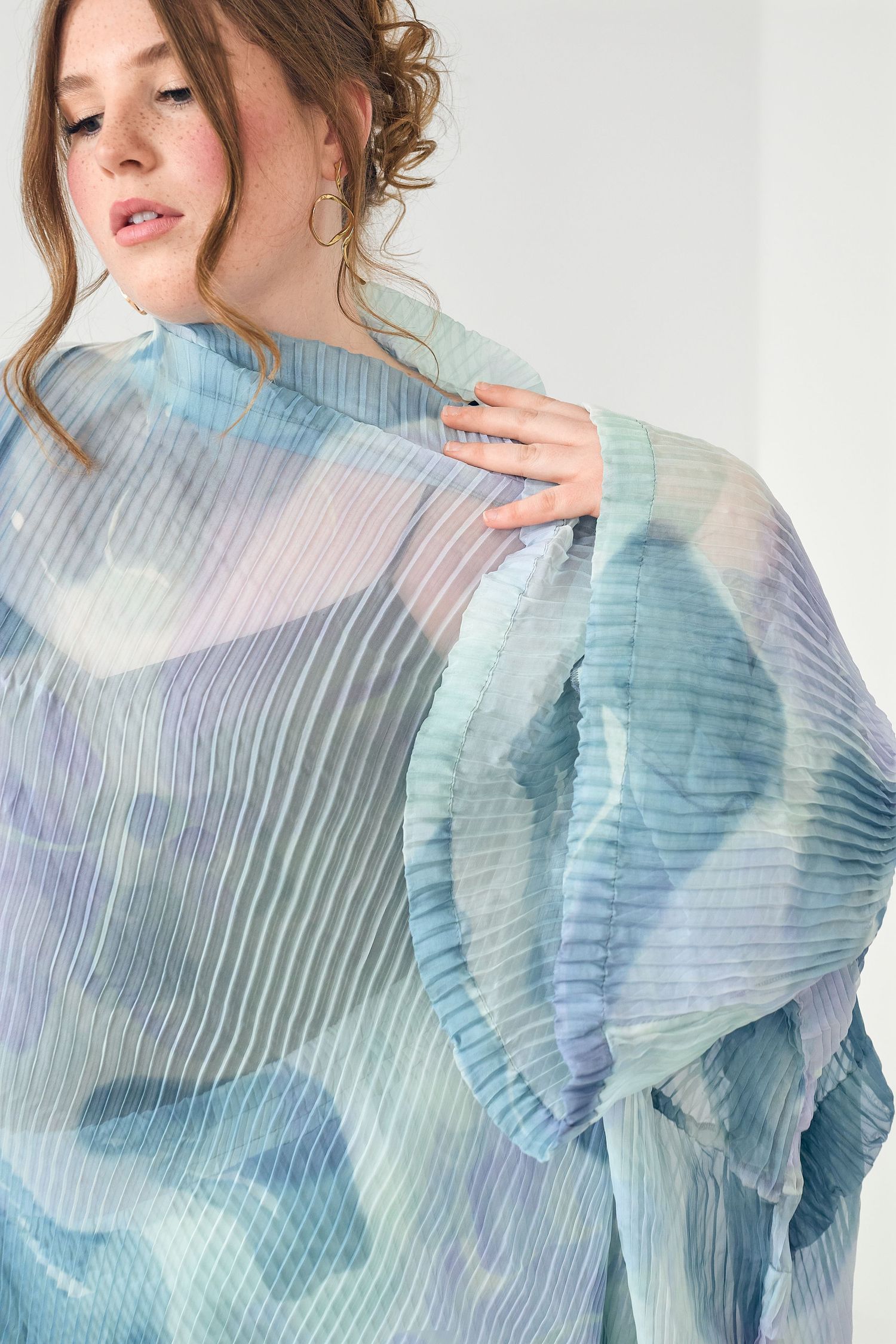 Woman wearing an oversized pleated blouse with soft blue and purple watercolor print, holding fabric up.