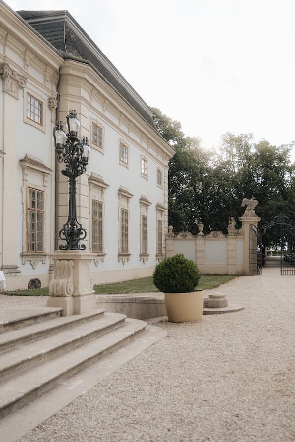 Elegant Baroque palace exterior with ornate lamppost, stone steps, trimmed shrub, and decorative gate on gravel path.